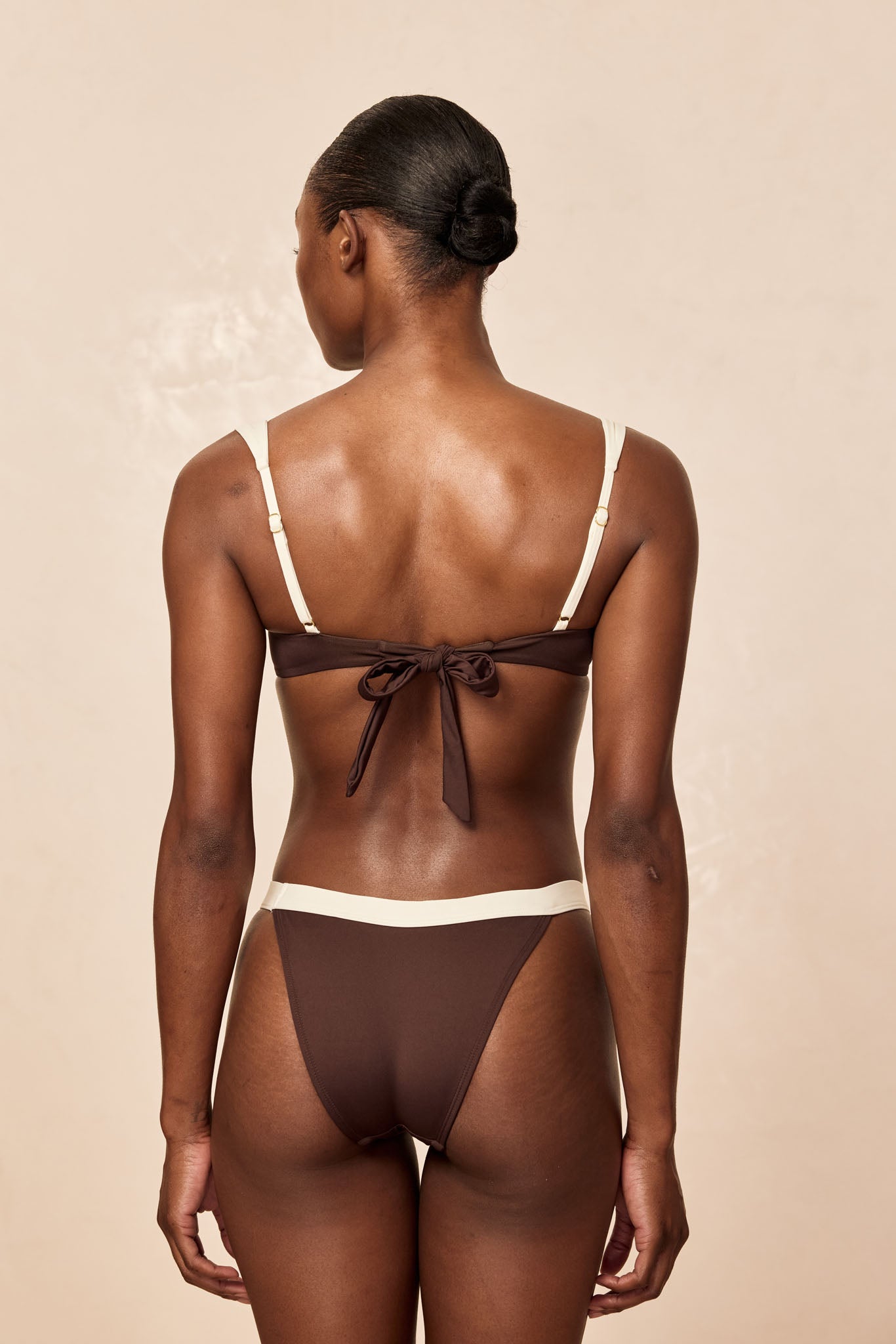Back view of a model wearing a brown bikini with cream straps and a tied back bow; hair in a neat low bun, beige background.