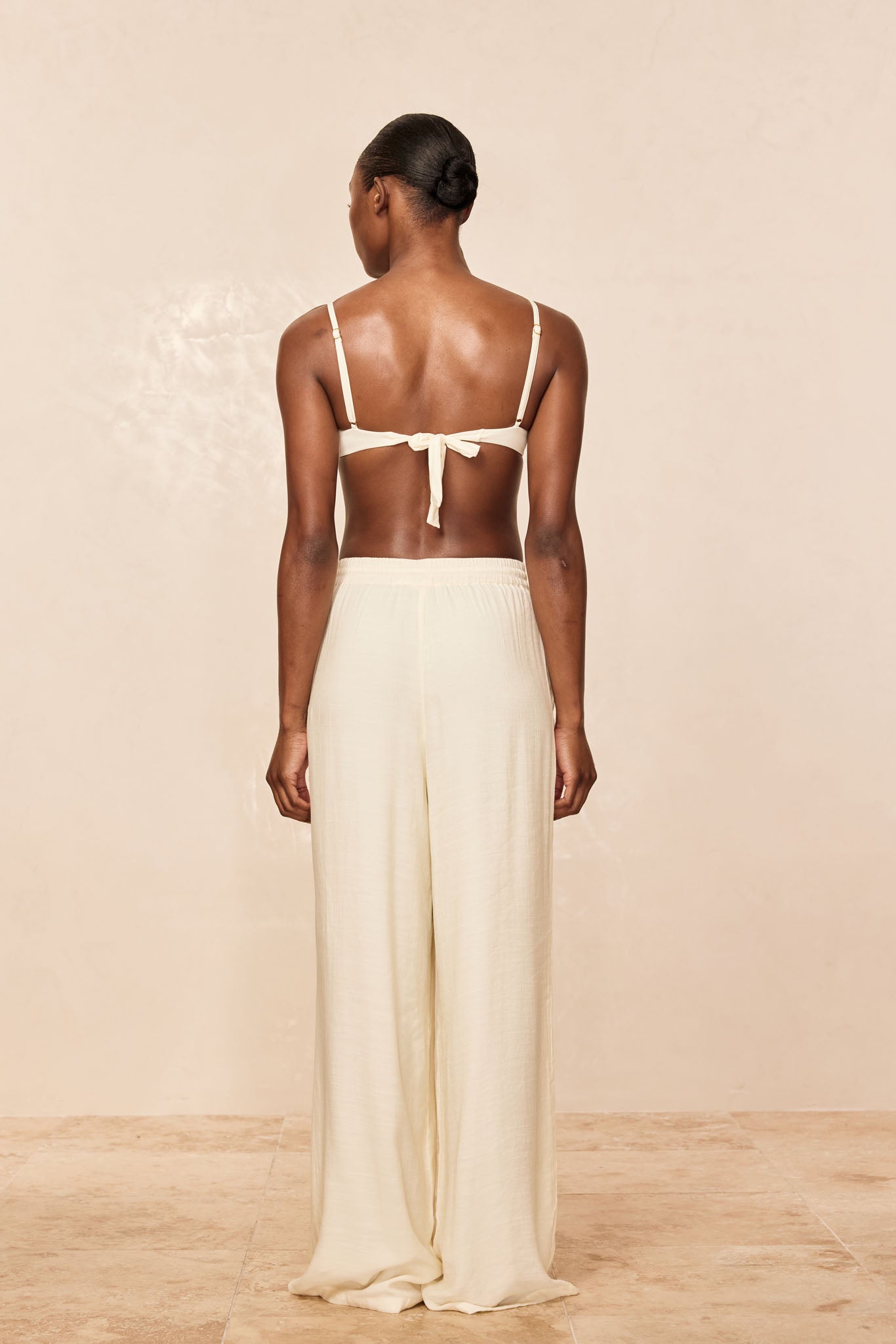 Back view of a model wearing a cream tie-back bikini top and high-waist, wide-leg cream pants on a beige background.
