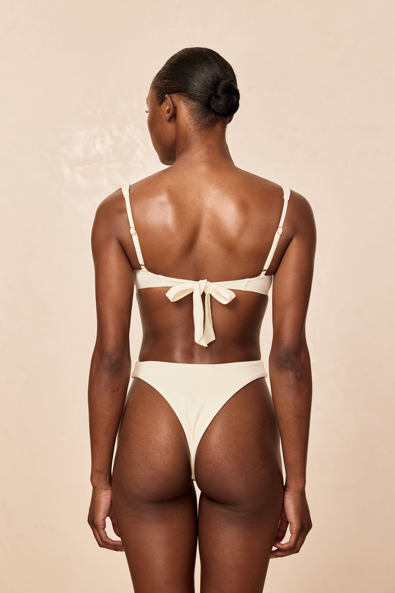 Woman shown from back wearing cream bikini with tied center-back bow and high-cut bottoms on neutral background.