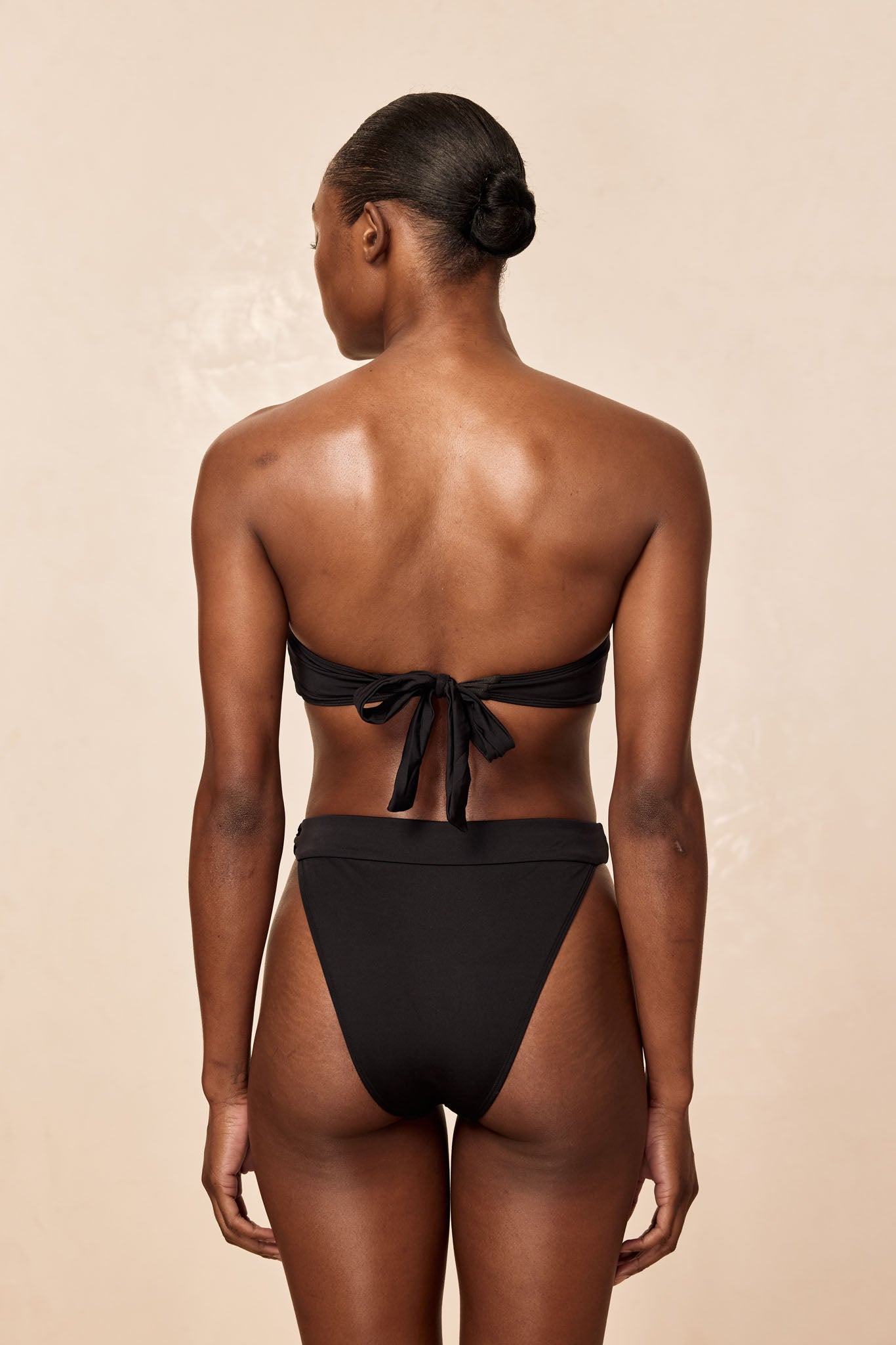 Back view of model wearing a black bandeau bikini top tied at center back and high-waist black bikini bottoms.