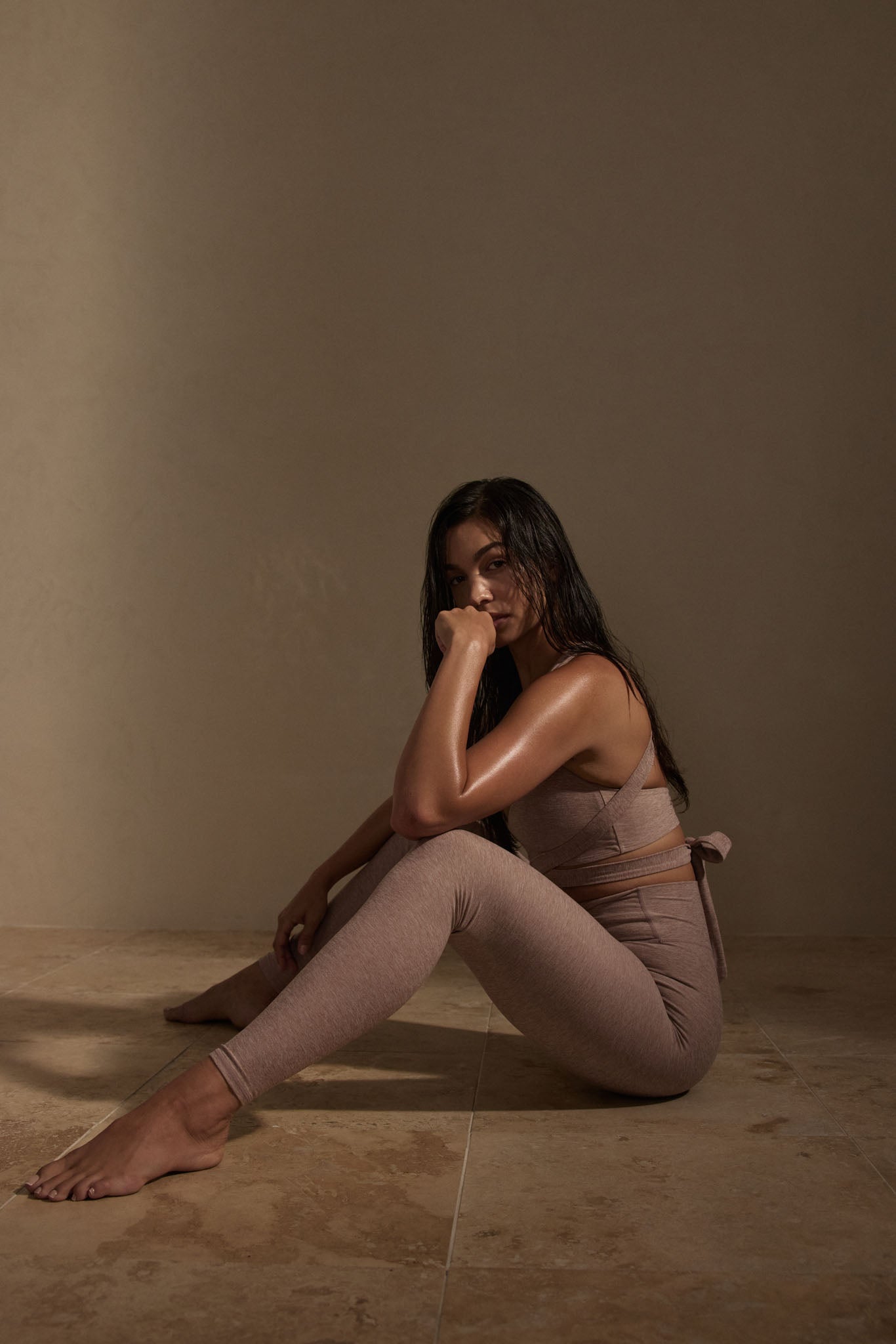 Female model seated on tile floor in mauve activewear, arms around knees, gazing toward camera.
