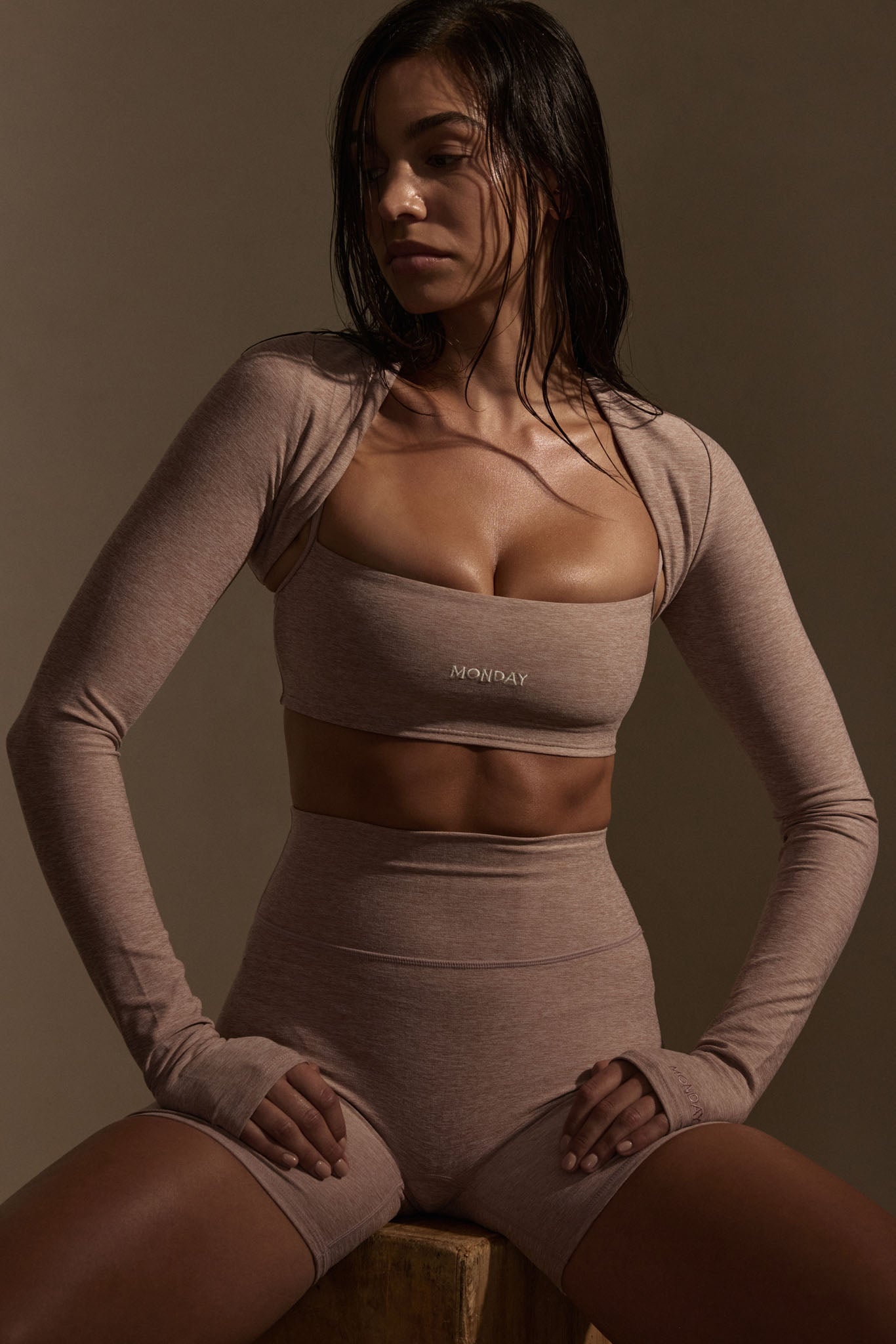 Seated woman in taupe workout set with bandeau top labeled MONDAY, shrug sleeves and high-waist shorts; hands resting on thighs.
