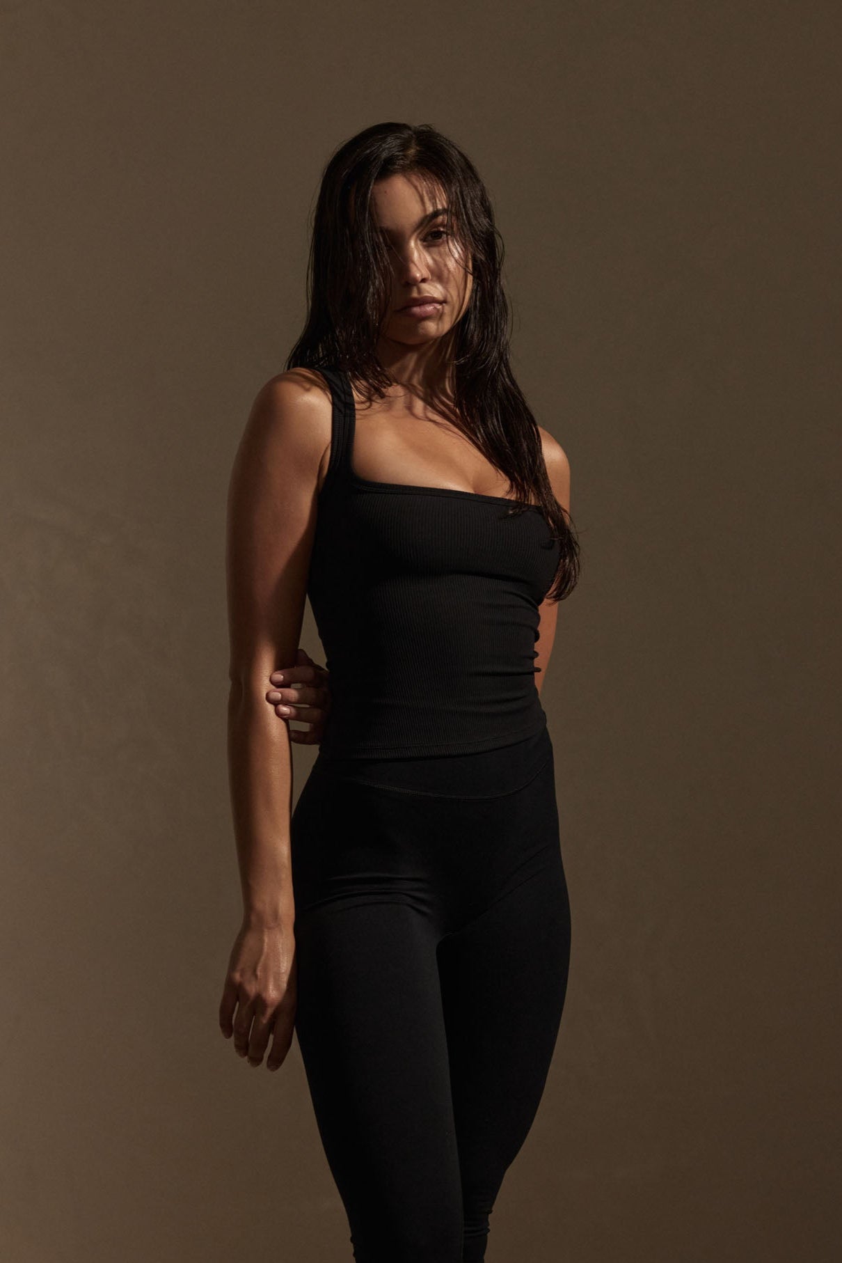 Woman standing in black tank top and leggings against a brown backdrop, wet hair and neutral expression, three-quarter pose.
