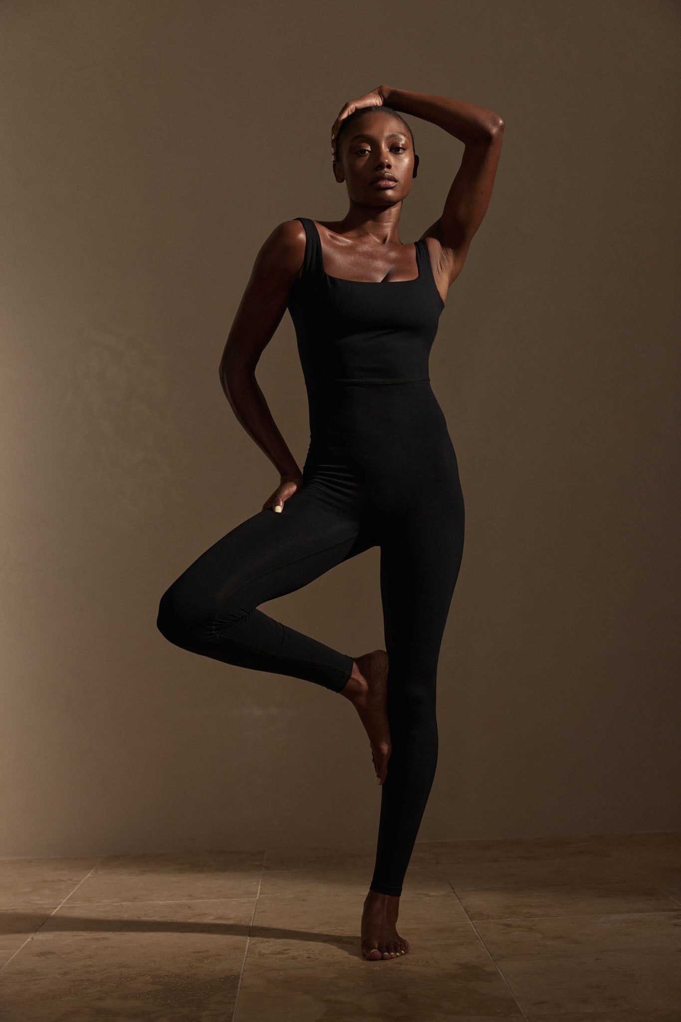 Woman in a black fitted sleeveless jumpsuit balancing on one leg, hand on head, against a warm brown studio backdrop.
