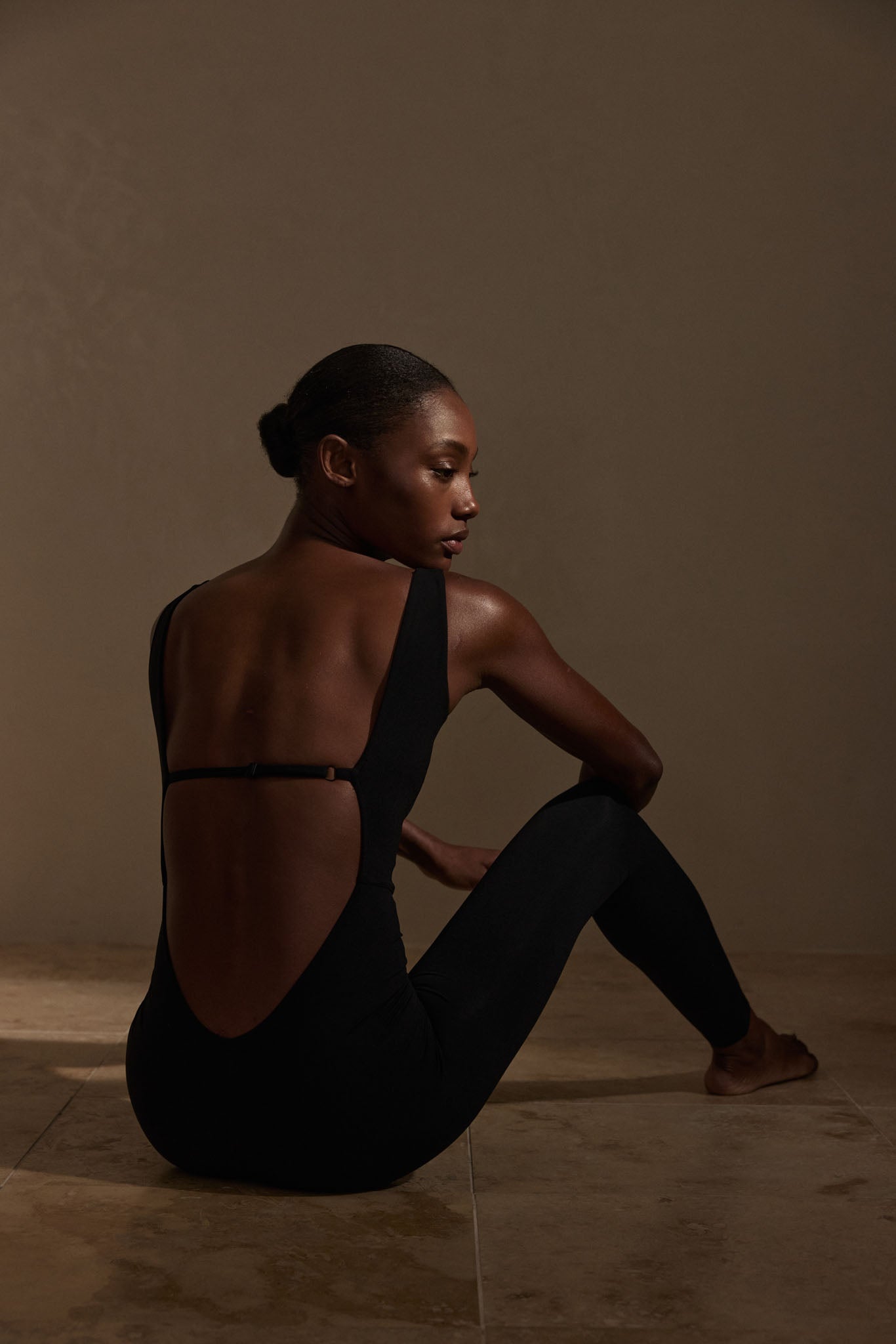 Woman seated on tiled floor, back exposed in a black backless leotard, turned profile under warm studio lighting.
