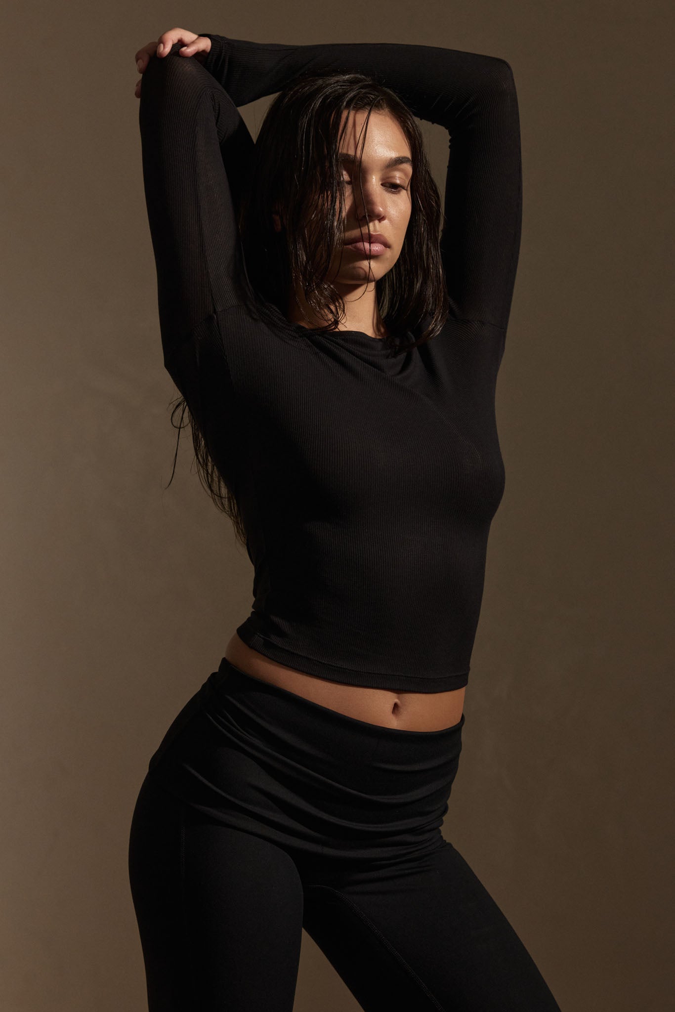 Woman in black long-sleeve top and leggings with arms raised behind her head, neutral expression, warm studio lighting