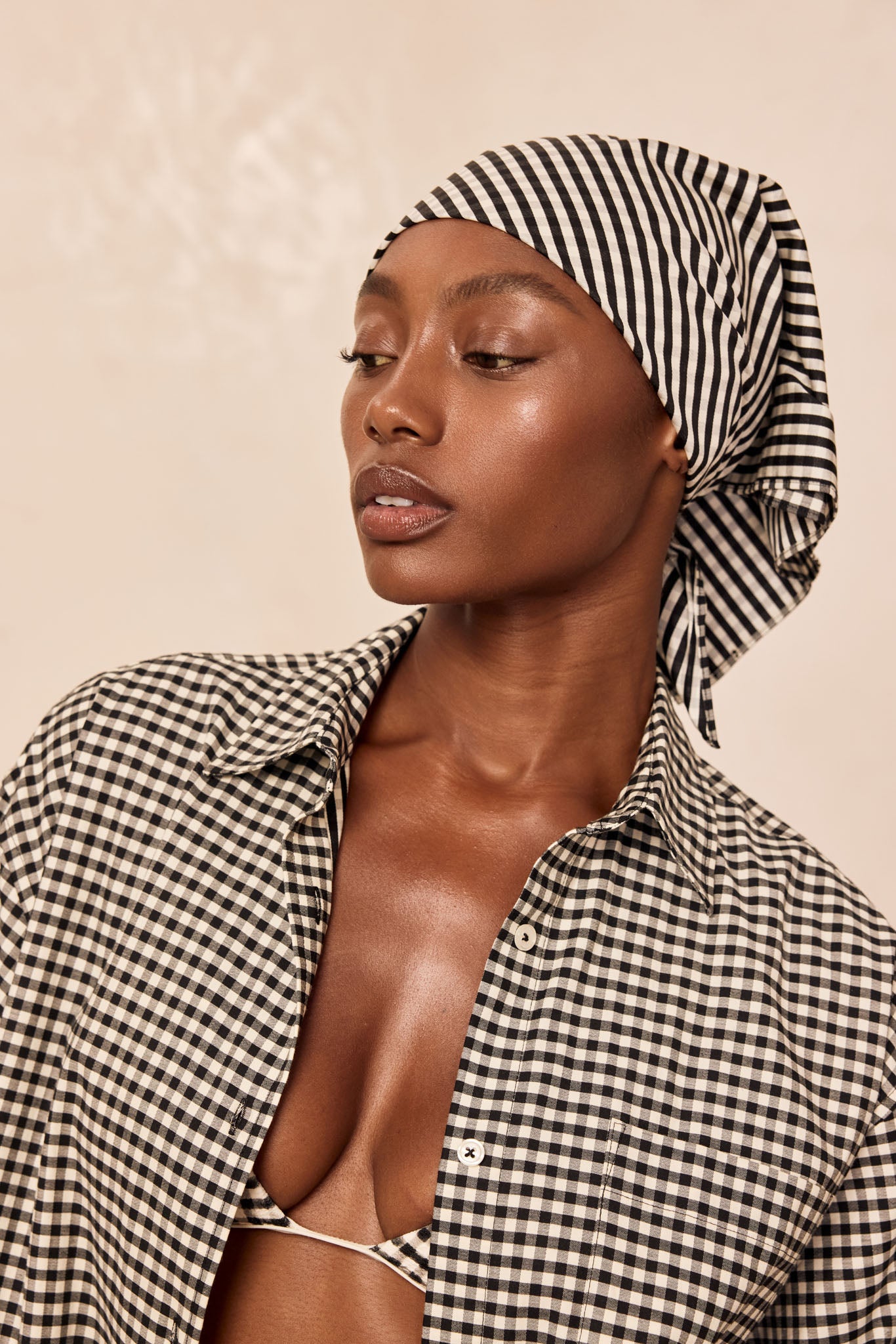 Close-up of woman in black-and-white gingham shirt and headscarf, gazing left, showing upper torso and collarbone.