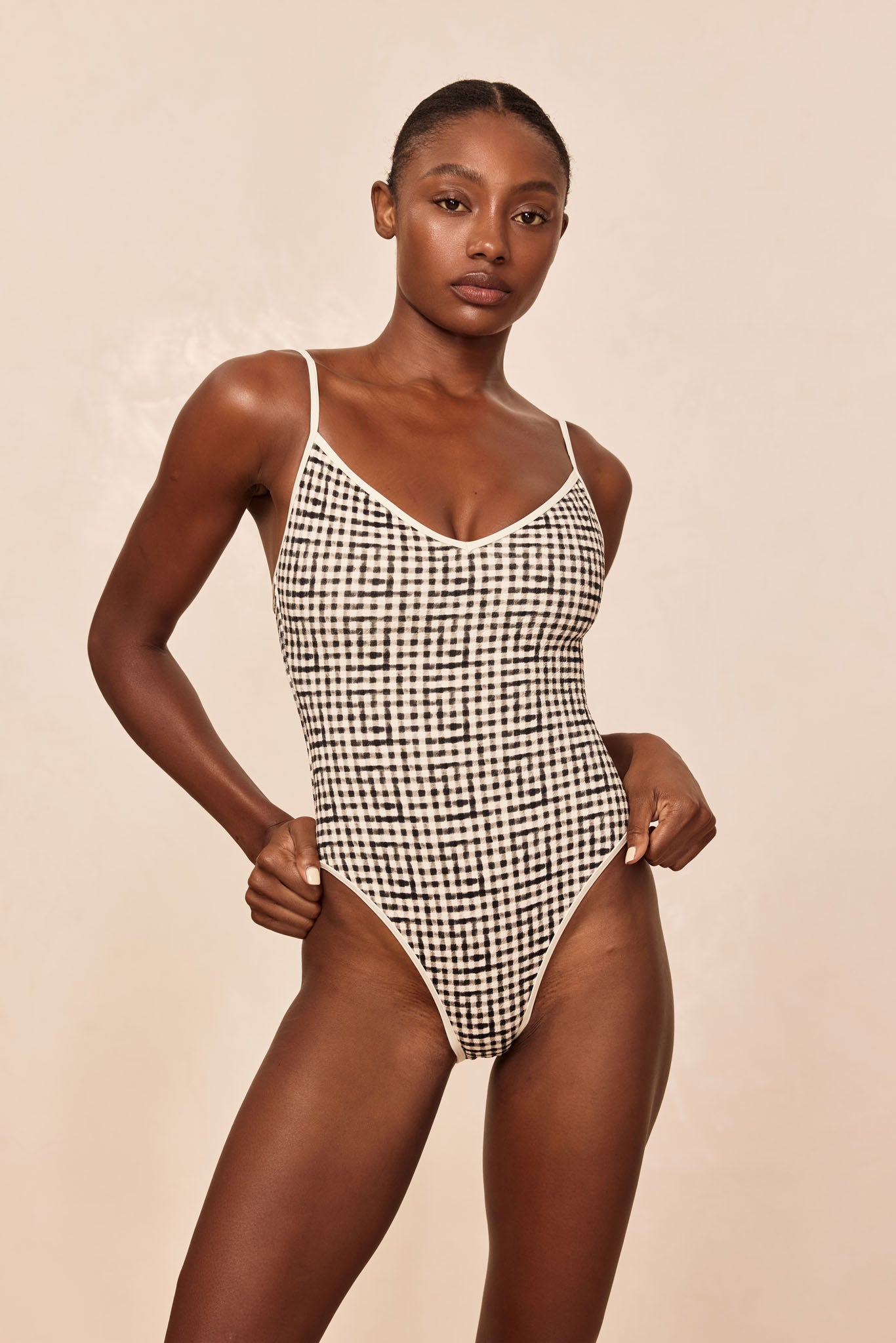 Model wearing black-and-white gingham one-piece swimsuit with thin straps and high-cut leg on beige studio background.