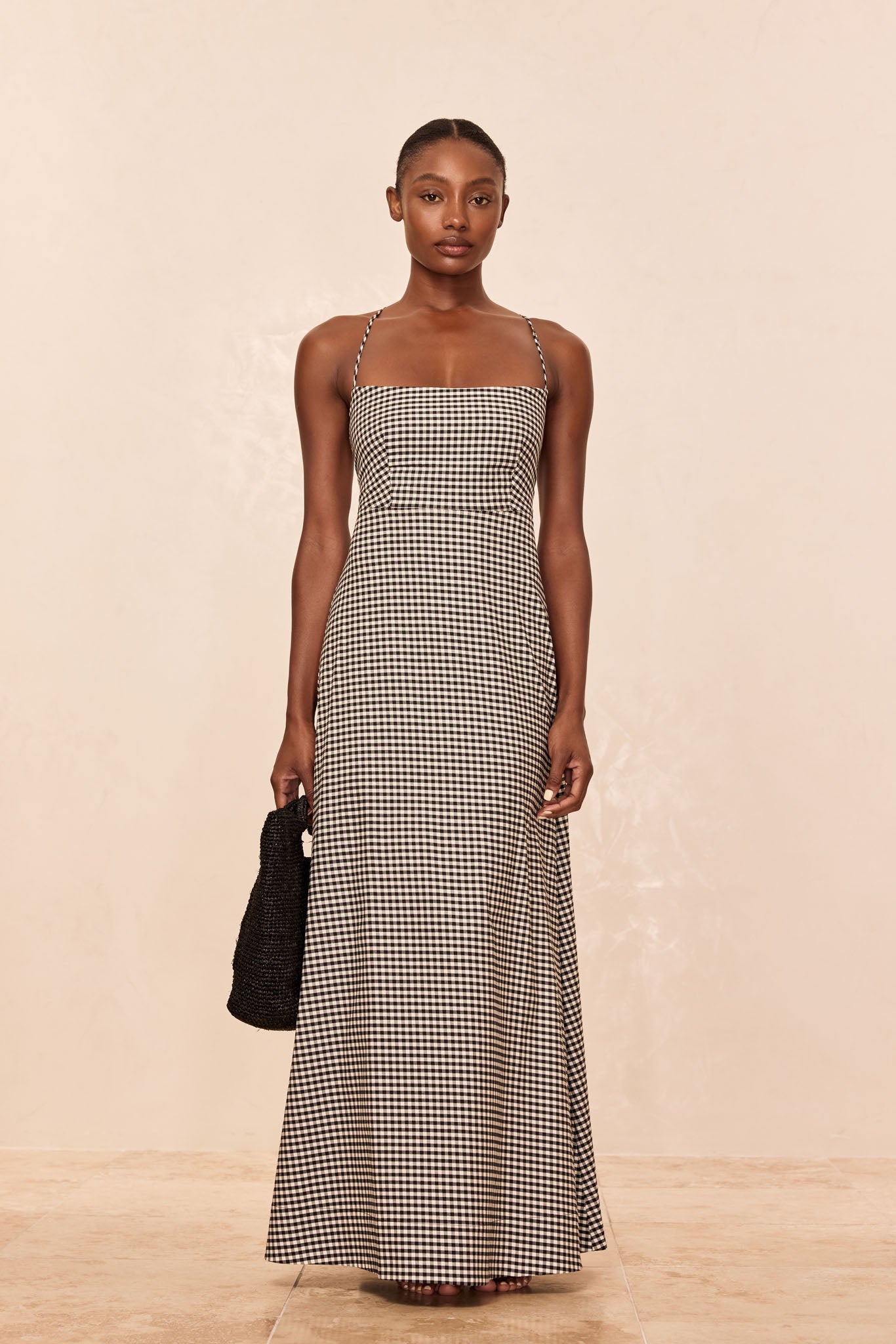 Model wearing a black-and-white gingham square-neck maxi dress with thin straps, holding a black woven tote.