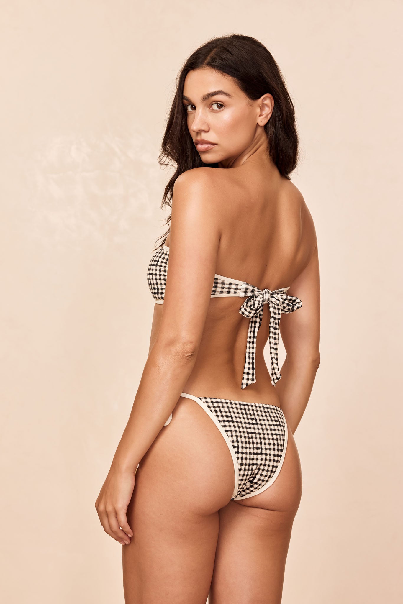 Woman seen from back, looking over her shoulder, wearing black-and-white gingham bandeau bikini with bow tied at back.