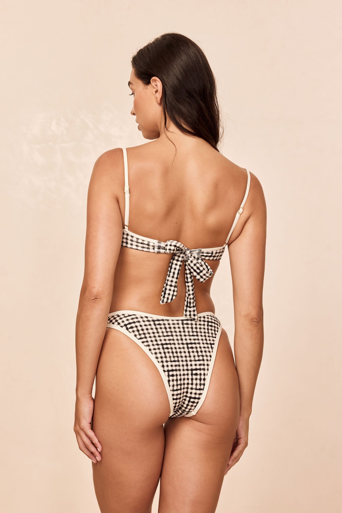 Back view of woman wearing high-cut black-and-white gingham tie-back bikini against beige background