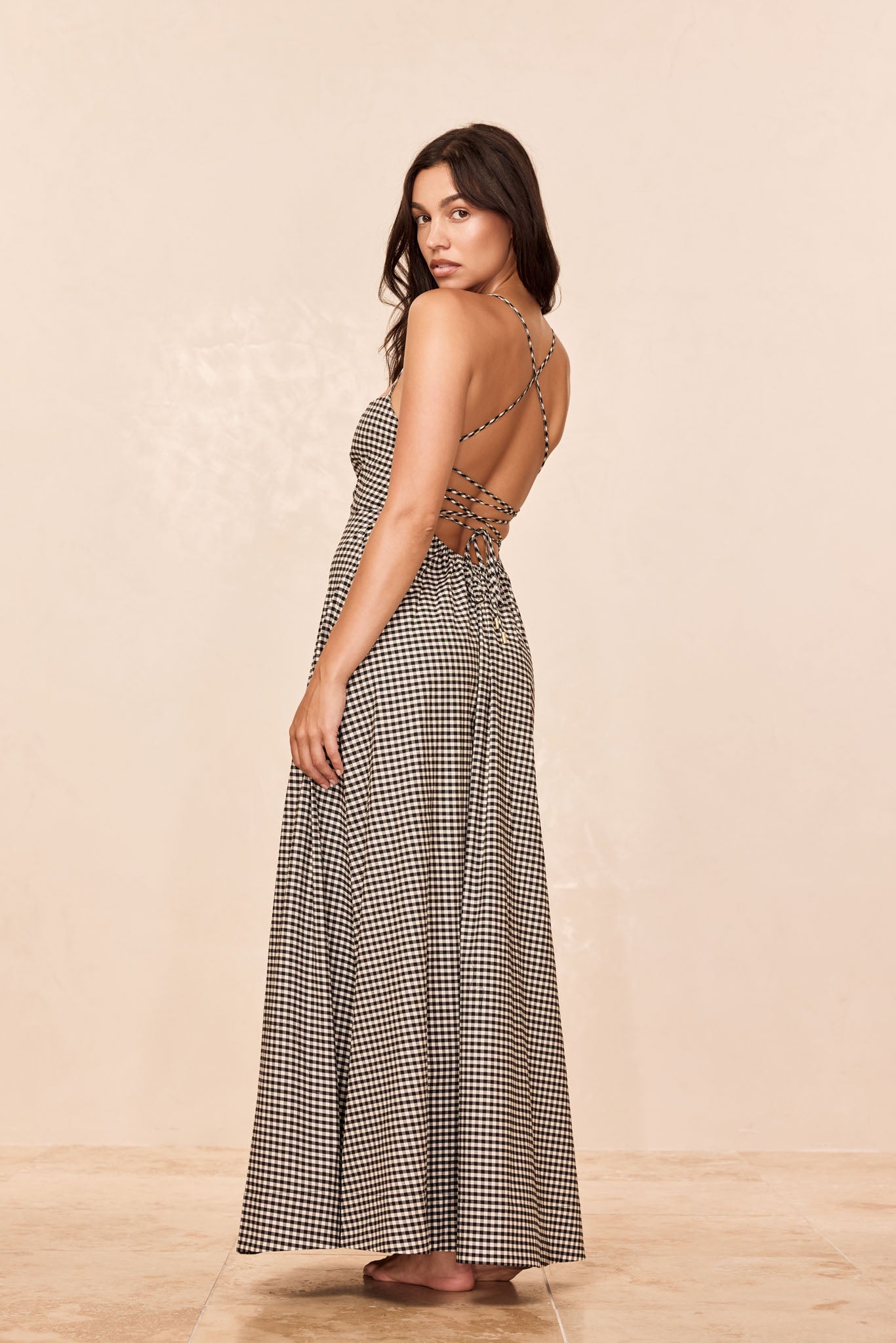 Model in a backless black-and-white gingham maxi dress with crisscross straps, looking over her shoulder, barefoot on tile.