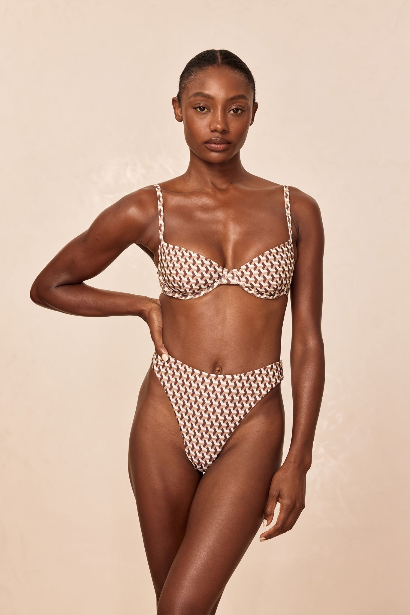 Woman modeling high-cut bikini set with brown-and-cream geometric print and underwire top against neutral background.