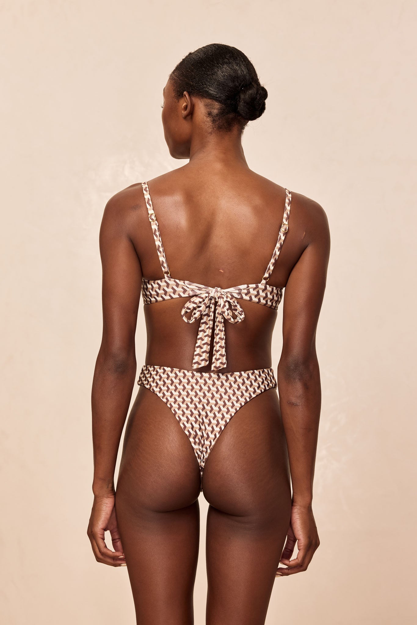 Rear view of a model wearing a brown-and-cream geometric-print bikini with a tied back top and high-cut bottoms.