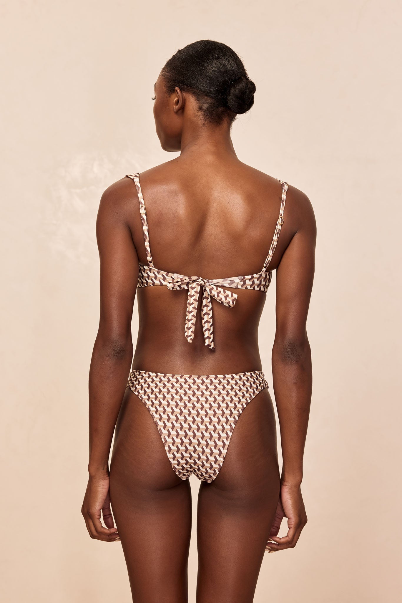Back view of model wearing brown-and-cream geometric-print bikini with tie-back top on neutral background.