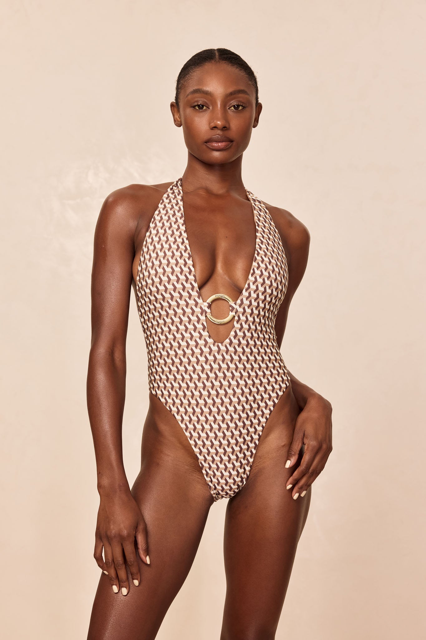 Woman modeling brown geometric-print halter one-piece swimsuit with deep V and gold circular clasp at center front