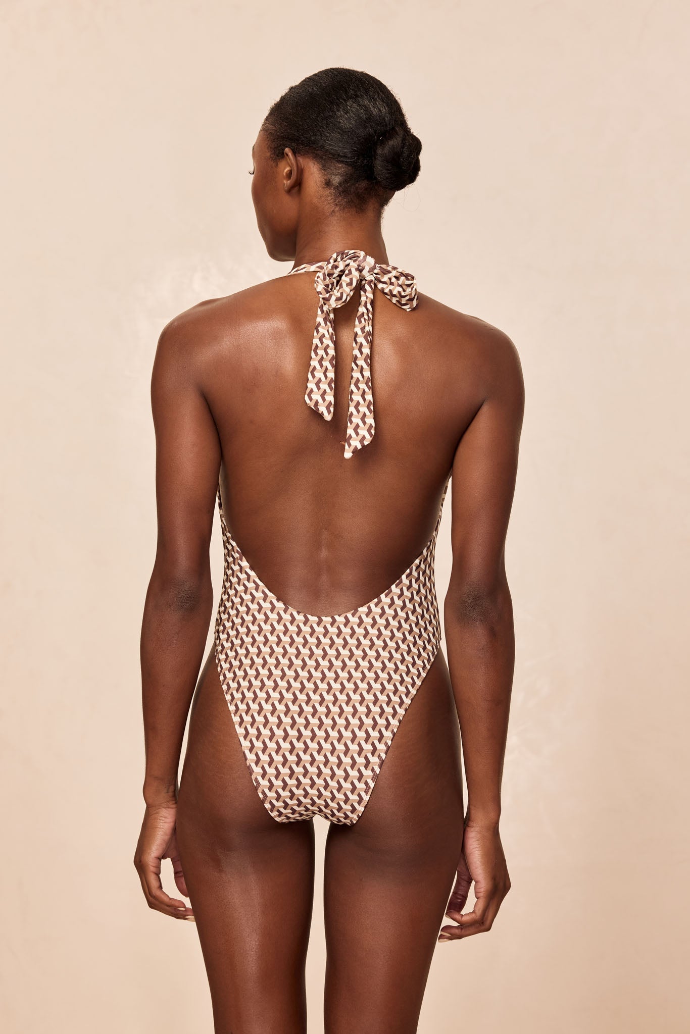 Rear view of a woman wearing a brown-and-cream patterned halter one-piece swimsuit with a tied bow at the neck.