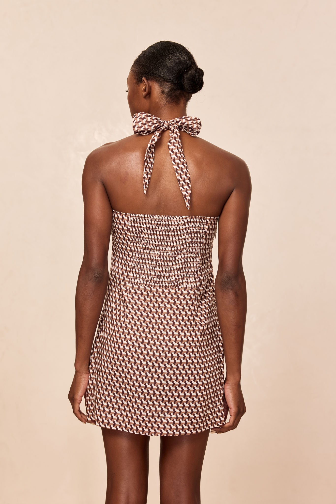 Back view of a woman wearing a brown-and-white geometric halter mini dress with smocked back and tied neck bow.