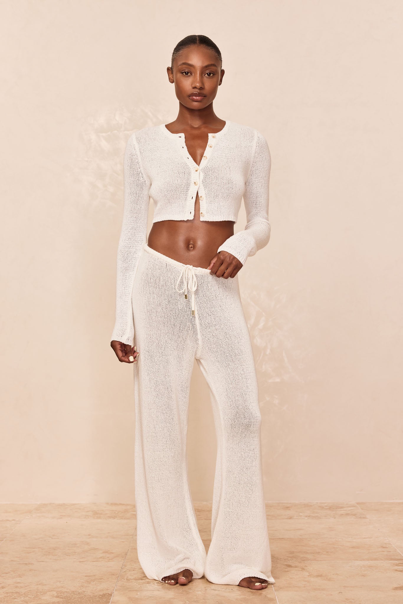 Model wearing white sheer knit long-sleeve cropped button-front cardigan and matching drawstring wide-leg pants.