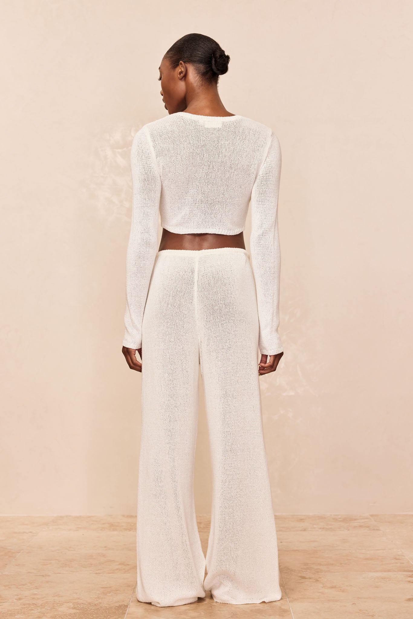 Back view of model wearing white sheer knit long-sleeve cropped top and high-waist wide-leg knit pants on beige backdrop