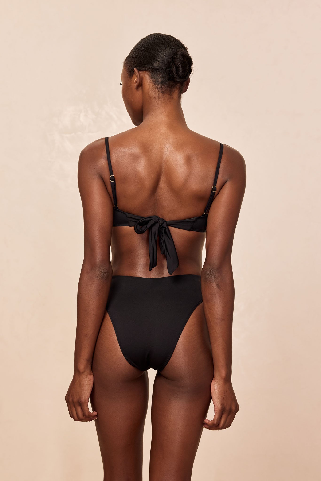 Model, back view, wearing black tie-back bikini top and high-rise black bikini bottoms on beige background.
