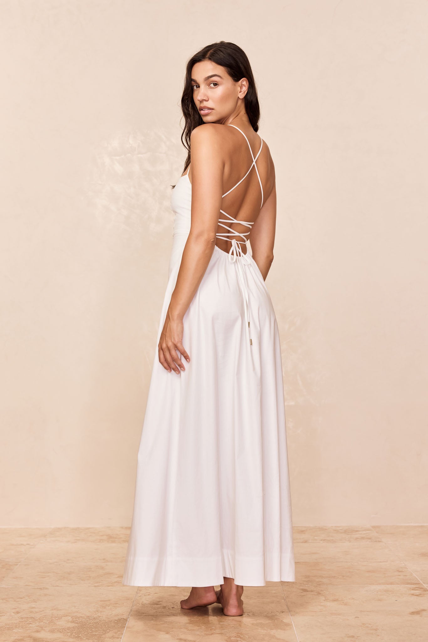 Woman looking over her shoulder wearing a floor-length white dress with crisscross back straps and waist tie