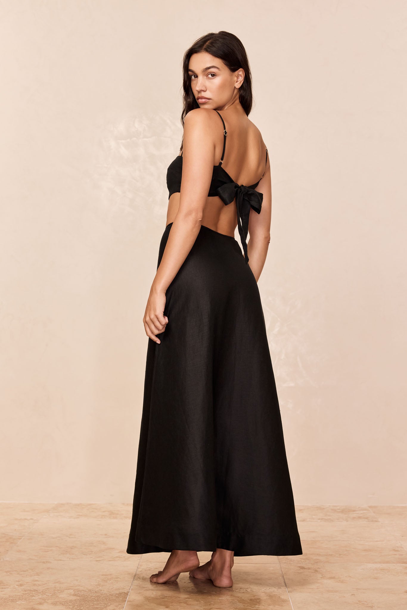 Back view of a barefoot woman wearing a black tie-back crop top and long skirt, looking over her shoulder.