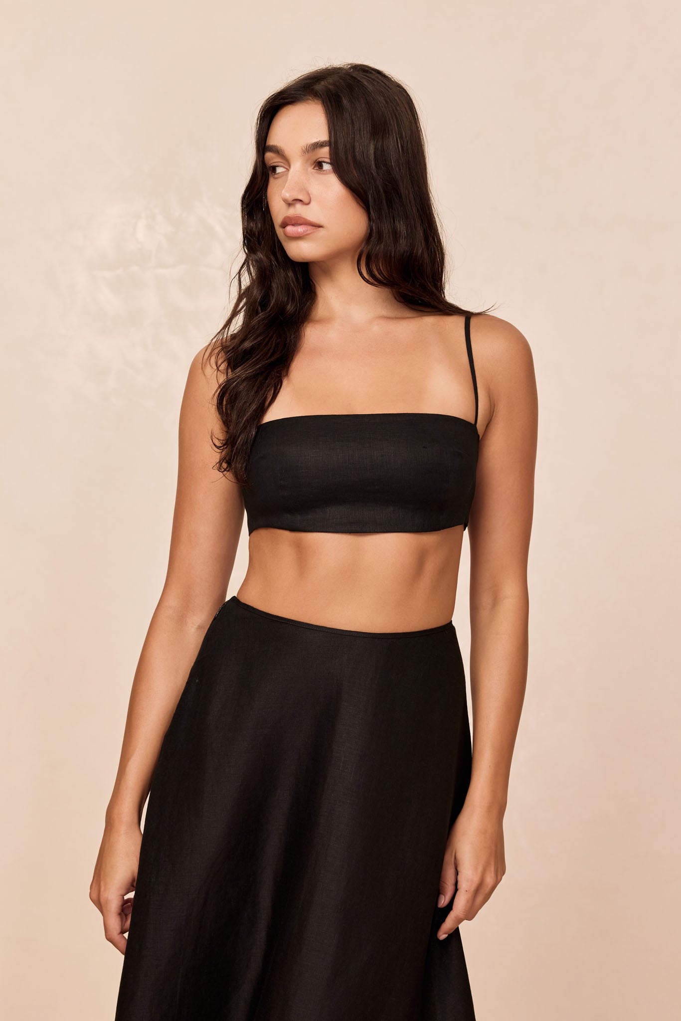 Woman wearing a black bandeau crop top and high-waist black skirt, standing against a beige background, looking left.