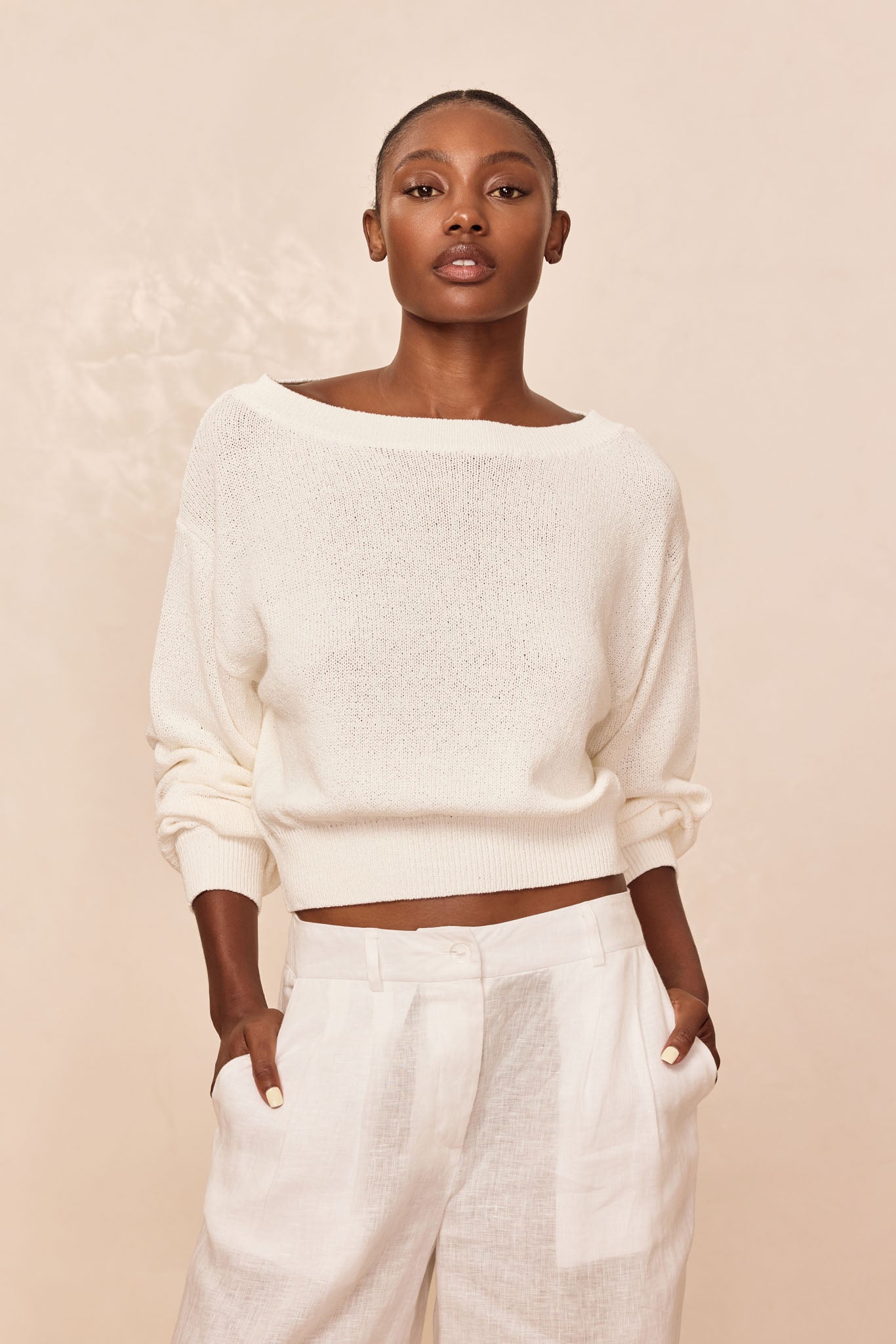 Model wearing an off-white boatneck knit sweater and high-waist white linen pants, hands in pockets against a neutral background