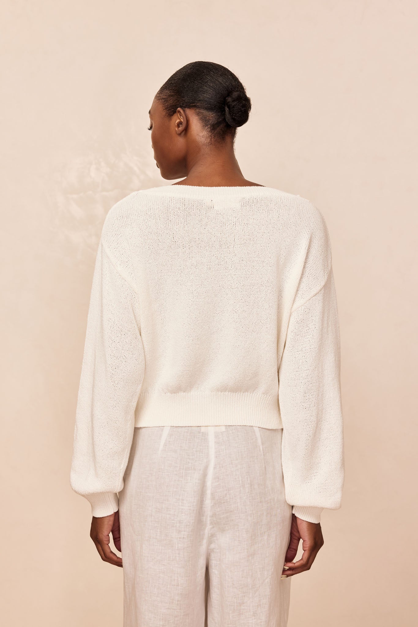 Rear view of person modeling a cream knit sweater and light linen pants, hair in a low bun against a neutral beige background.