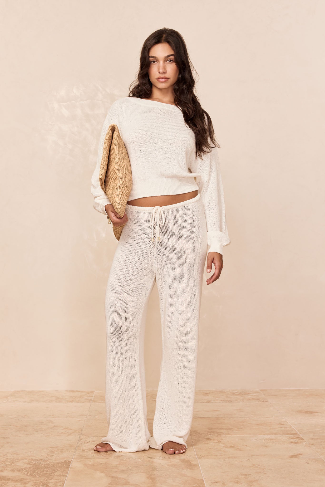 Model wearing cream knit long-sleeve sweater and drawstring wide-leg pants, holding a woven clutch, on beige tile floor