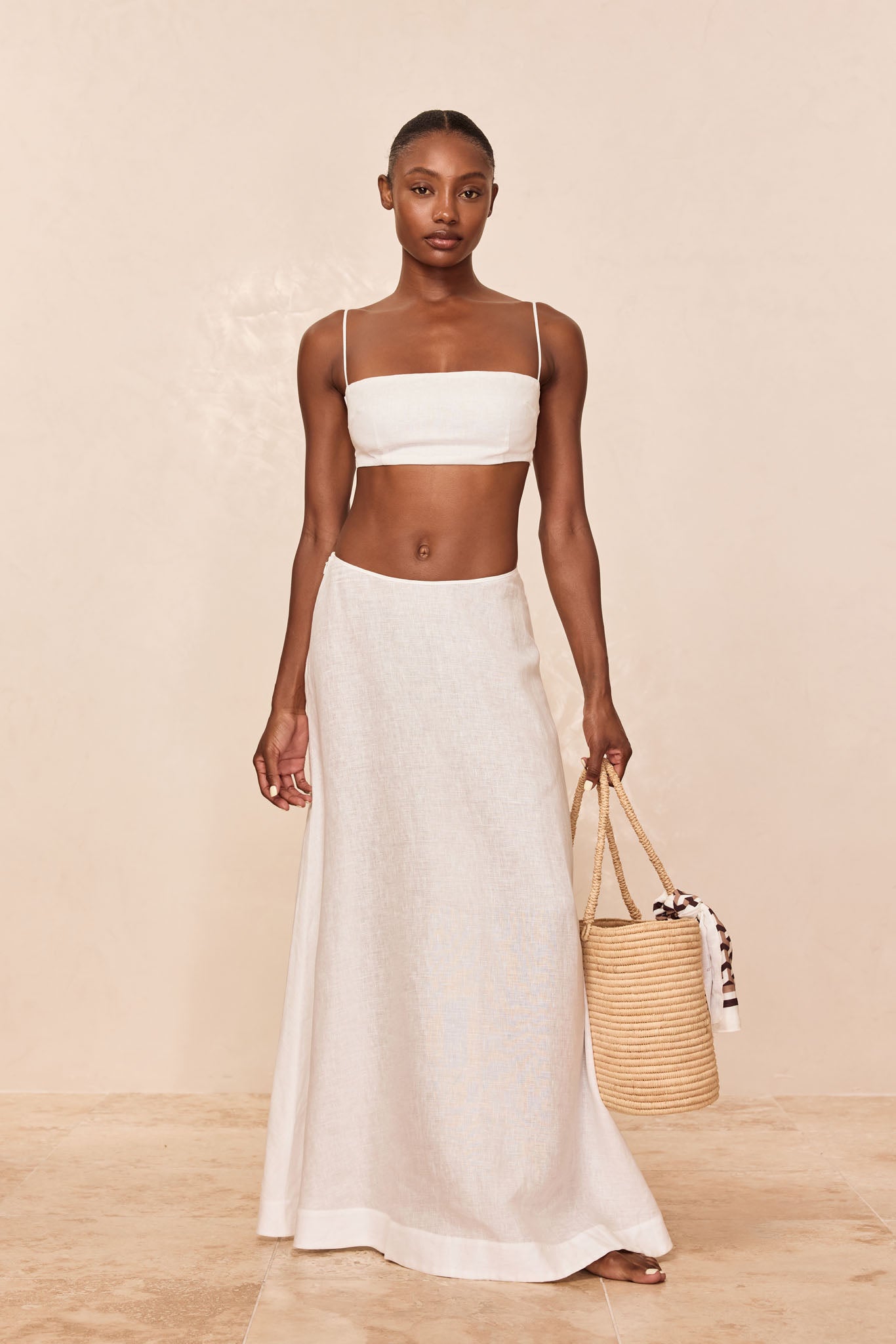 Model wearing white linen bandeau top and long A-line linen skirt, holding a woven straw bucket tote with knotted scarf.