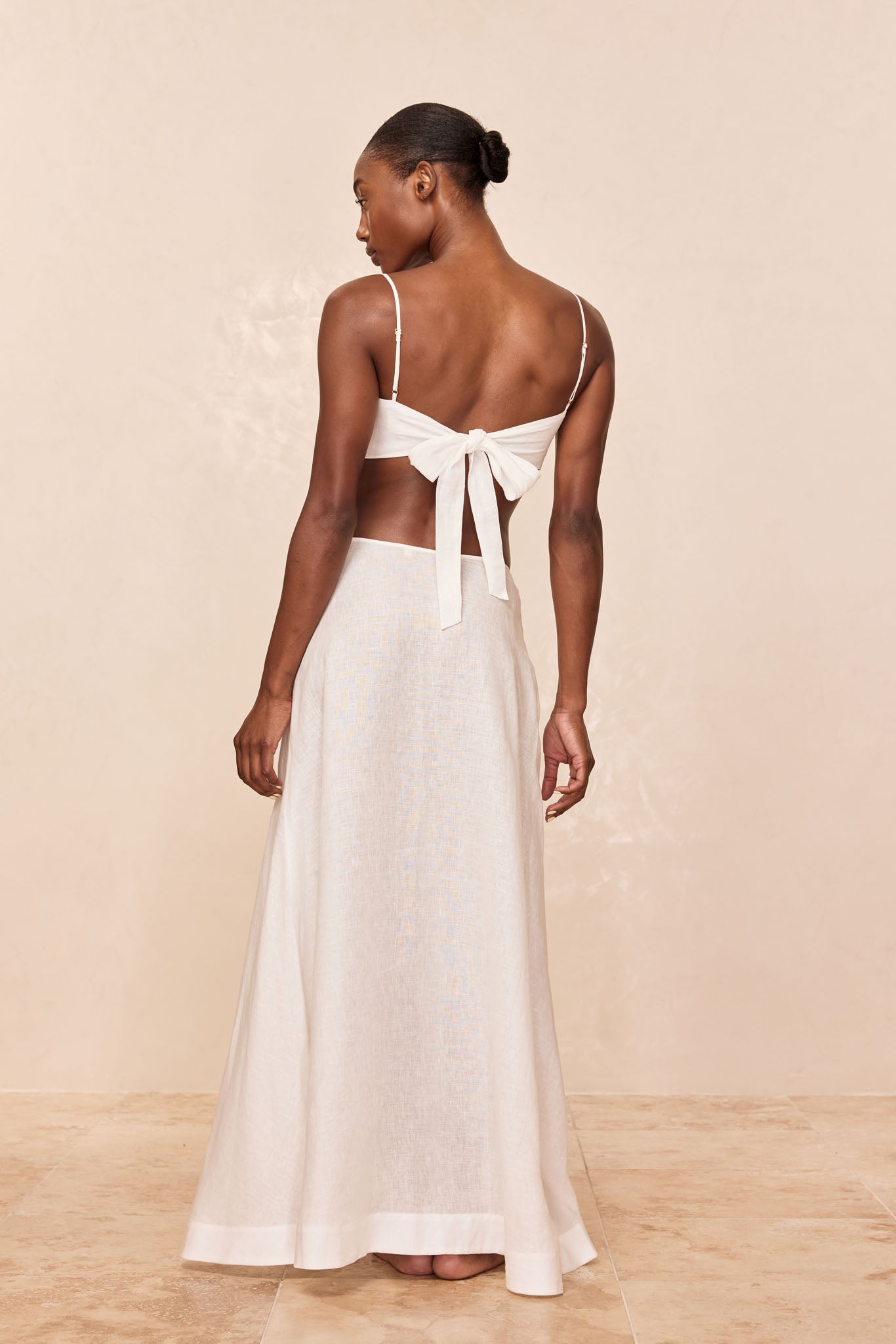 Back view of model wearing a white tie-back crop top and ankle-length white linen skirt against a neutral background.