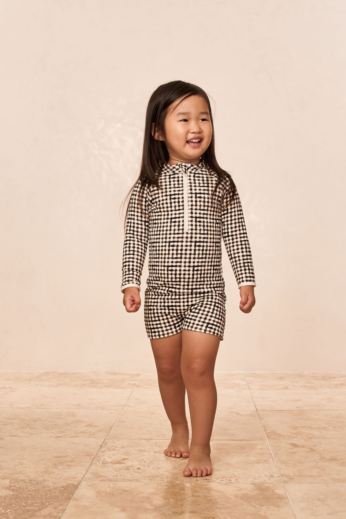 Smiling young girl walking barefoot in a black-and-white checkered long-sleeve romper with front zipper.