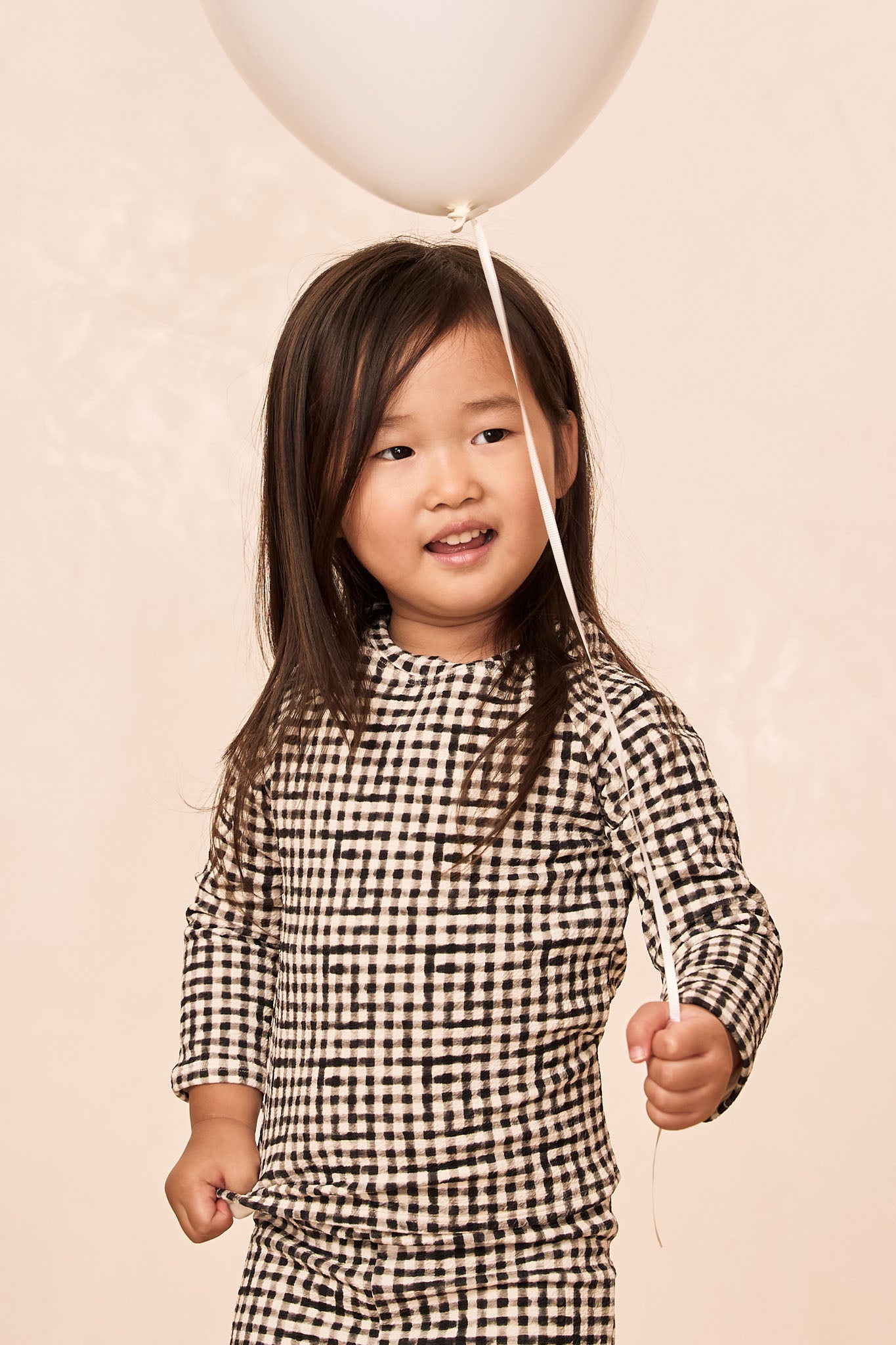 Young child in black-and-white gingham outfit holding a white balloon by its ribbon, smiling and looking to the right.