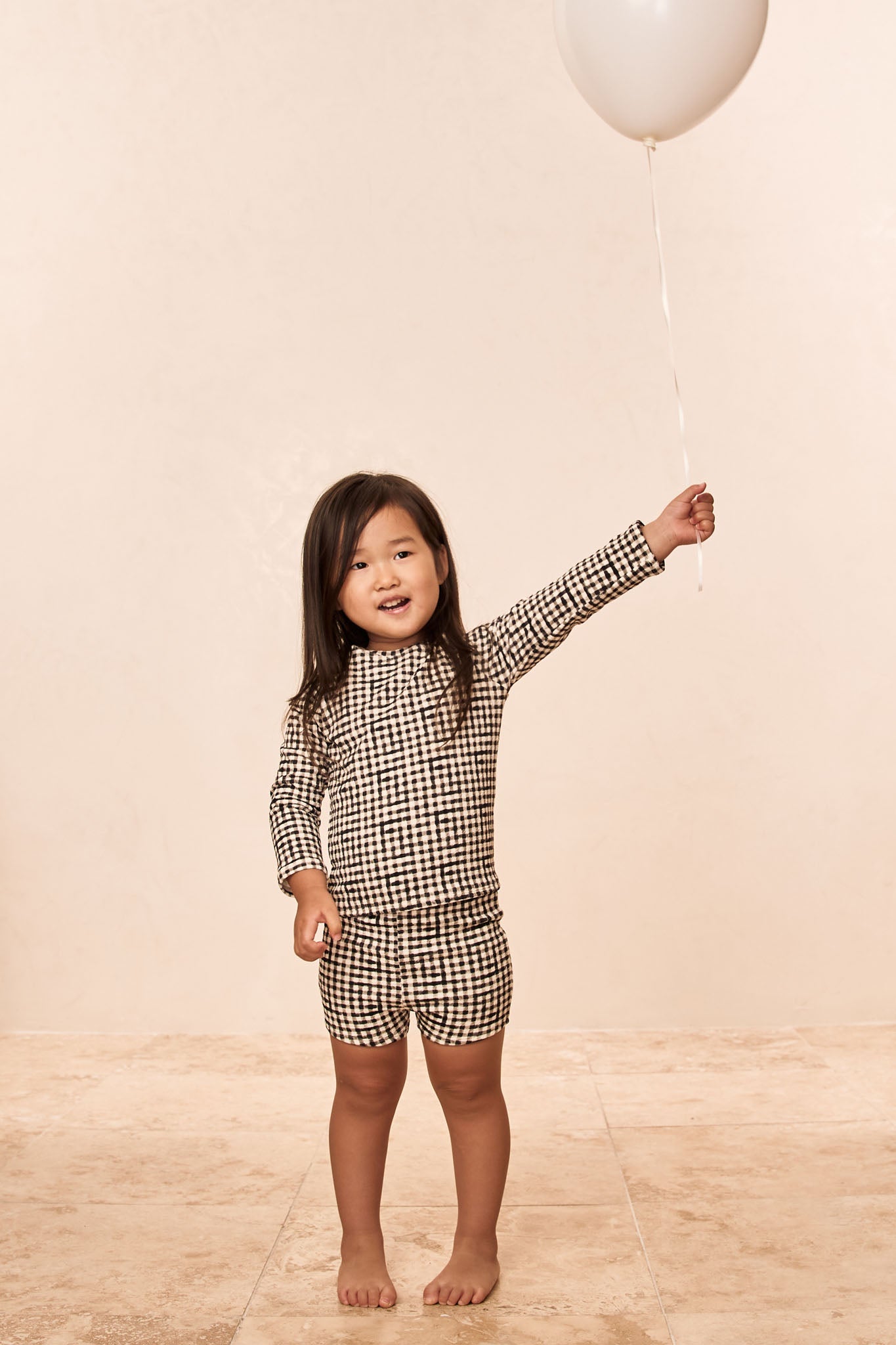 Young girl standing barefoot on tile holding a white balloon by its string, wearing a black-and-white check outfit, smiling.