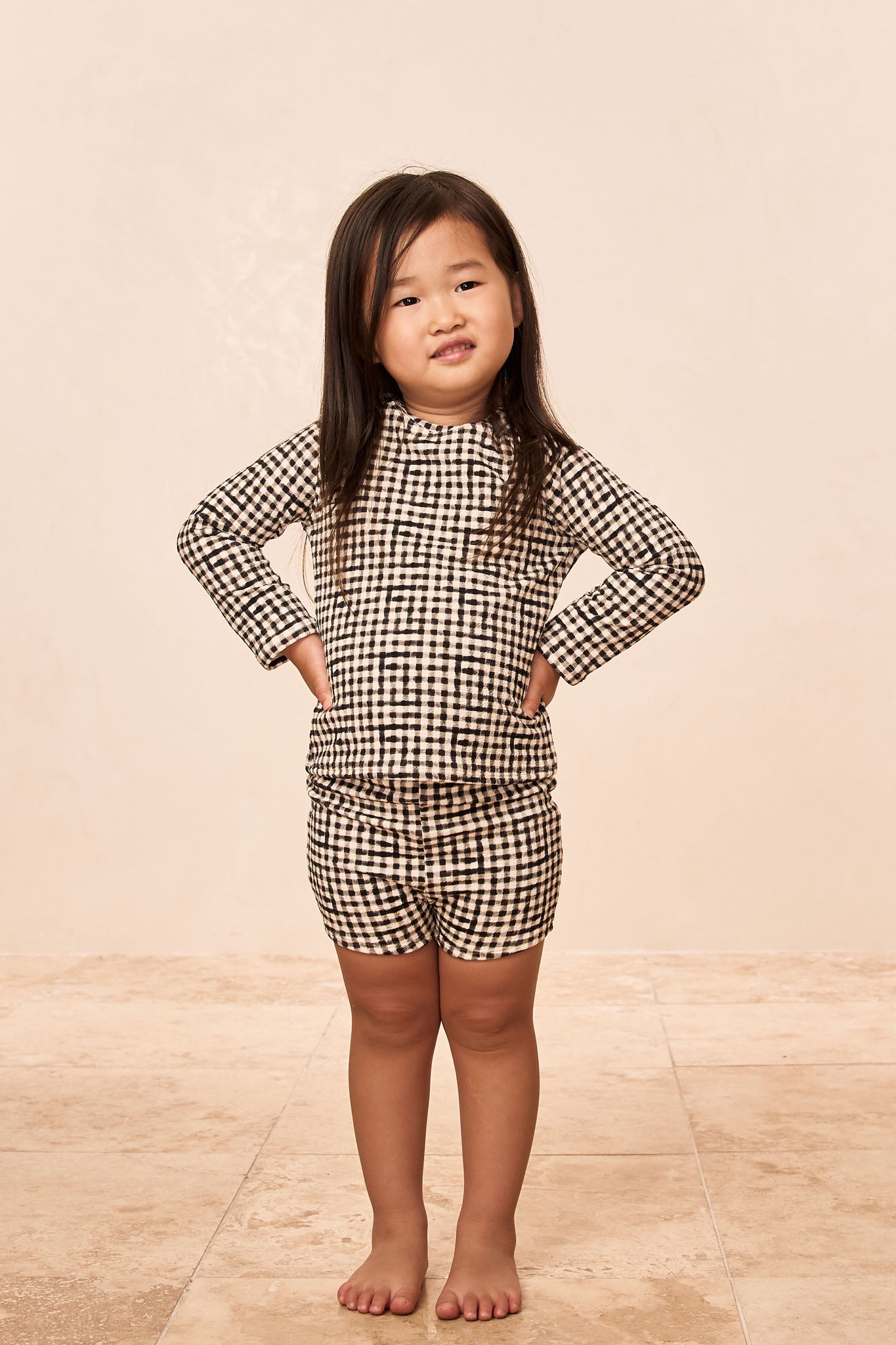 Child standing barefoot on tiled floor, hands on hips, wearing black-and-white gingham long-sleeve top and matching shorts.