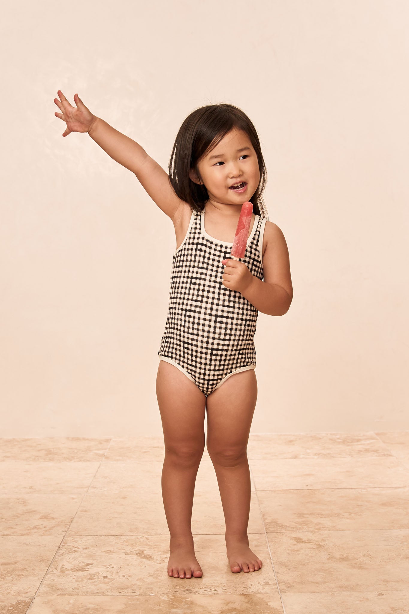 Toddler in black-and-white gingham swimsuit holding a red popsicle, raising one arm and smiling.