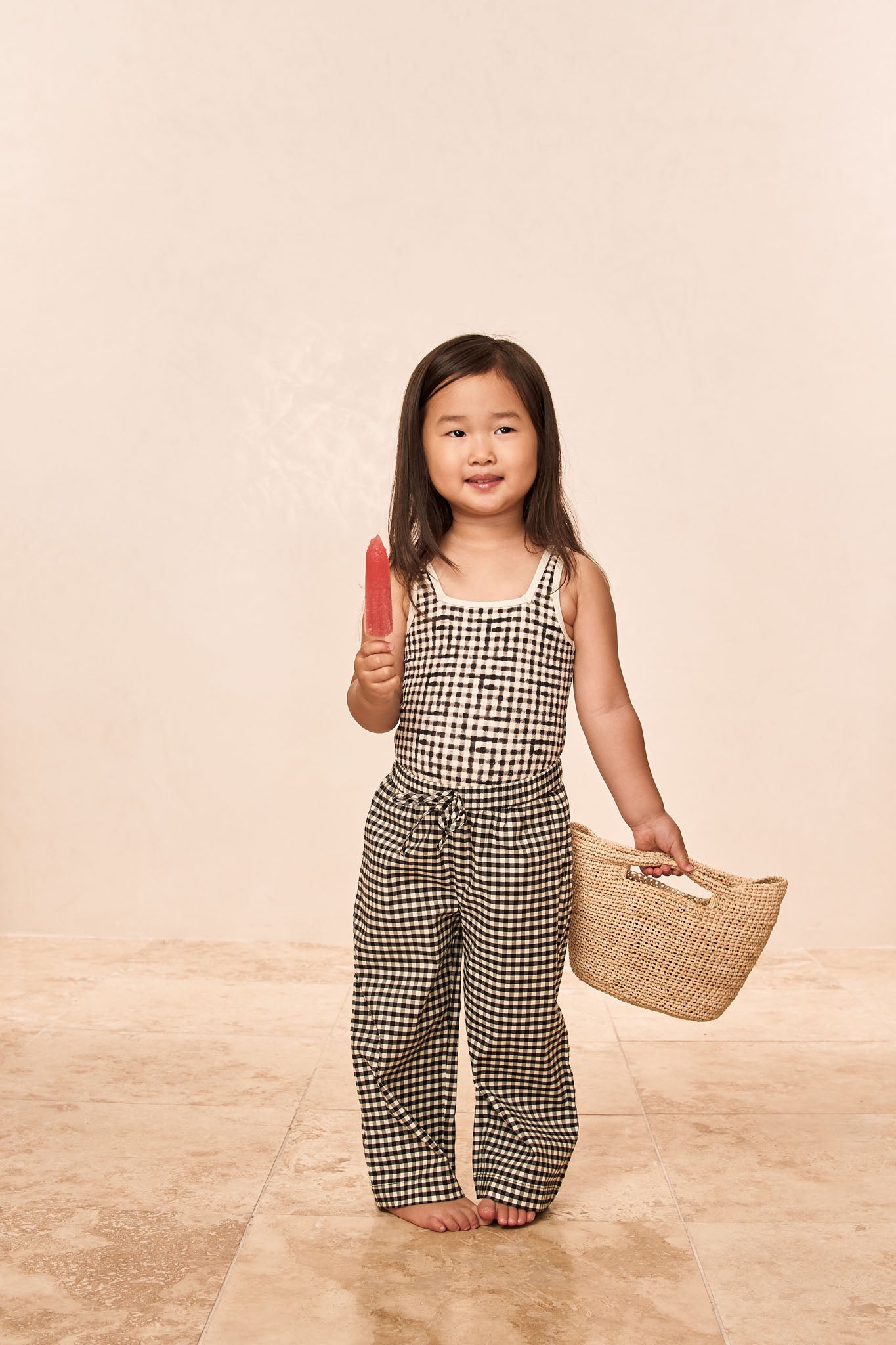 Young girl in black-and-white gingham top and pants holding a red popsicle and woven basket, smiling.