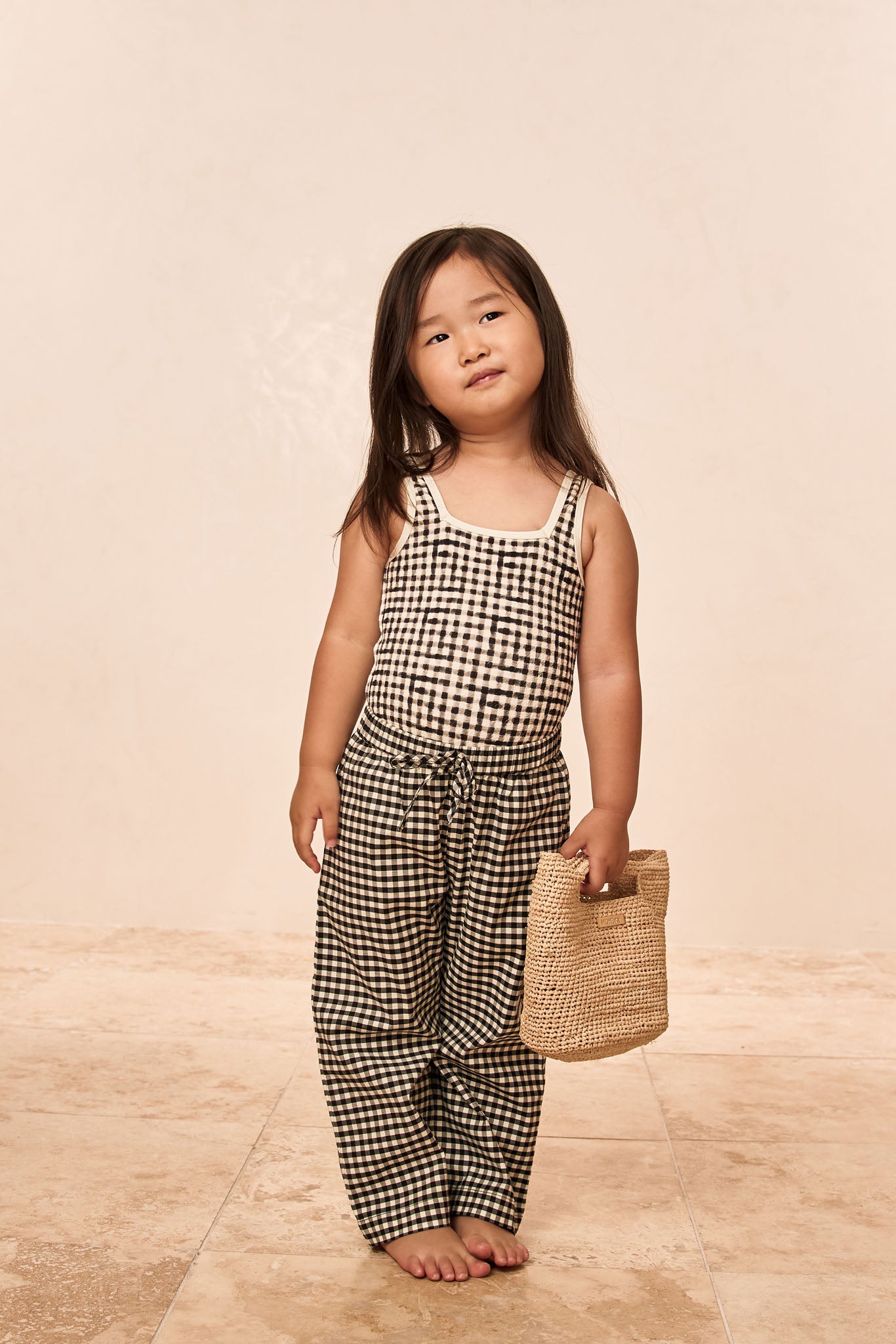 Young girl standing barefoot in a black-and-white gingham tank and pants, holding a woven straw tote, neutral studio background.