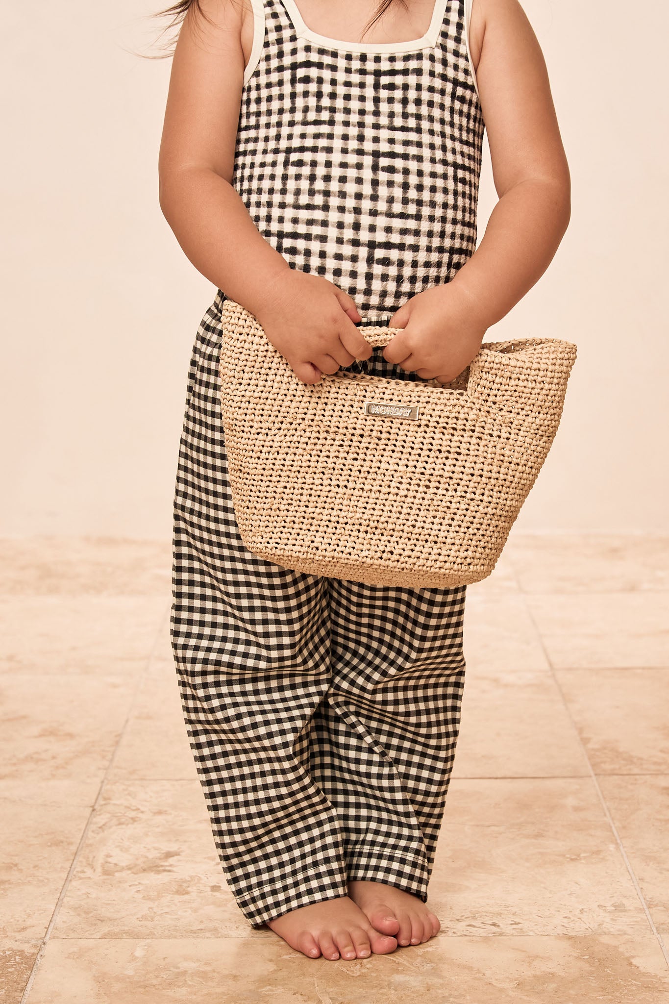Child barefoot in sleeveless black-and-white gingham top and pants holding a woven straw handbag at waist