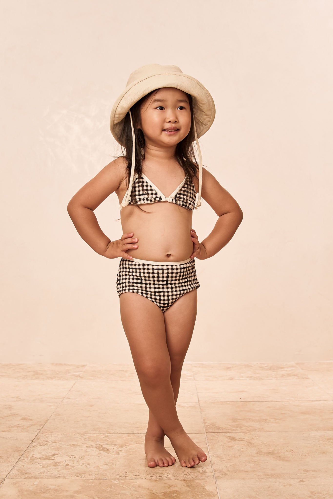 Young girl standing with hands on hips wearing gingham two-piece swimsuit and bucket hat on tiled floor.
