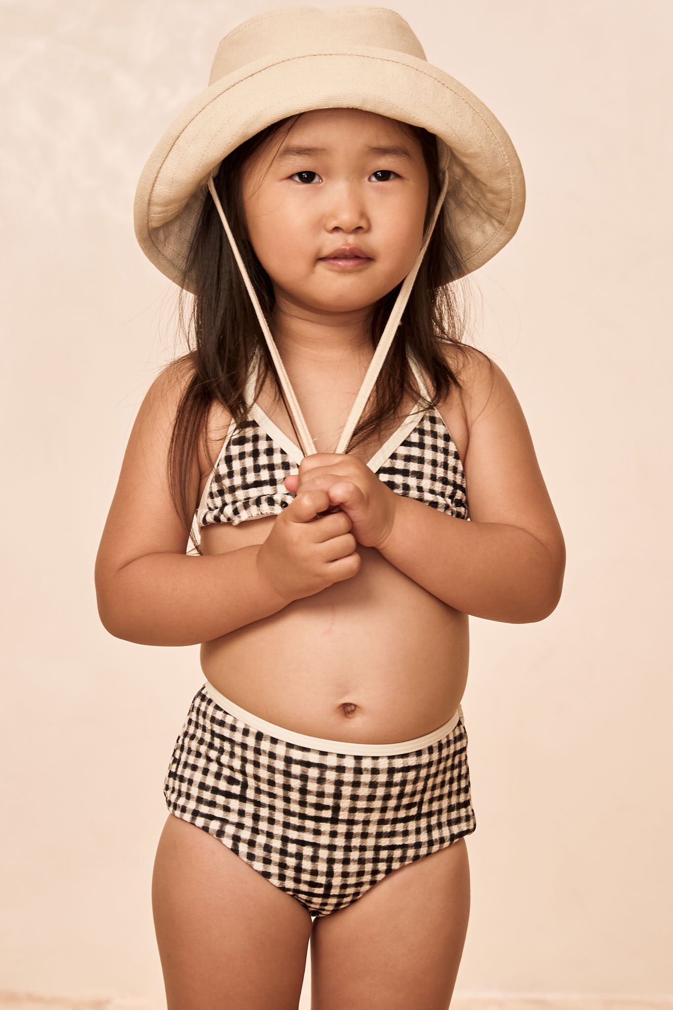 Young girl in a beige bucket hat and black-and-white gingham bikini, clasping the hat's cords against a neutral backdrop.