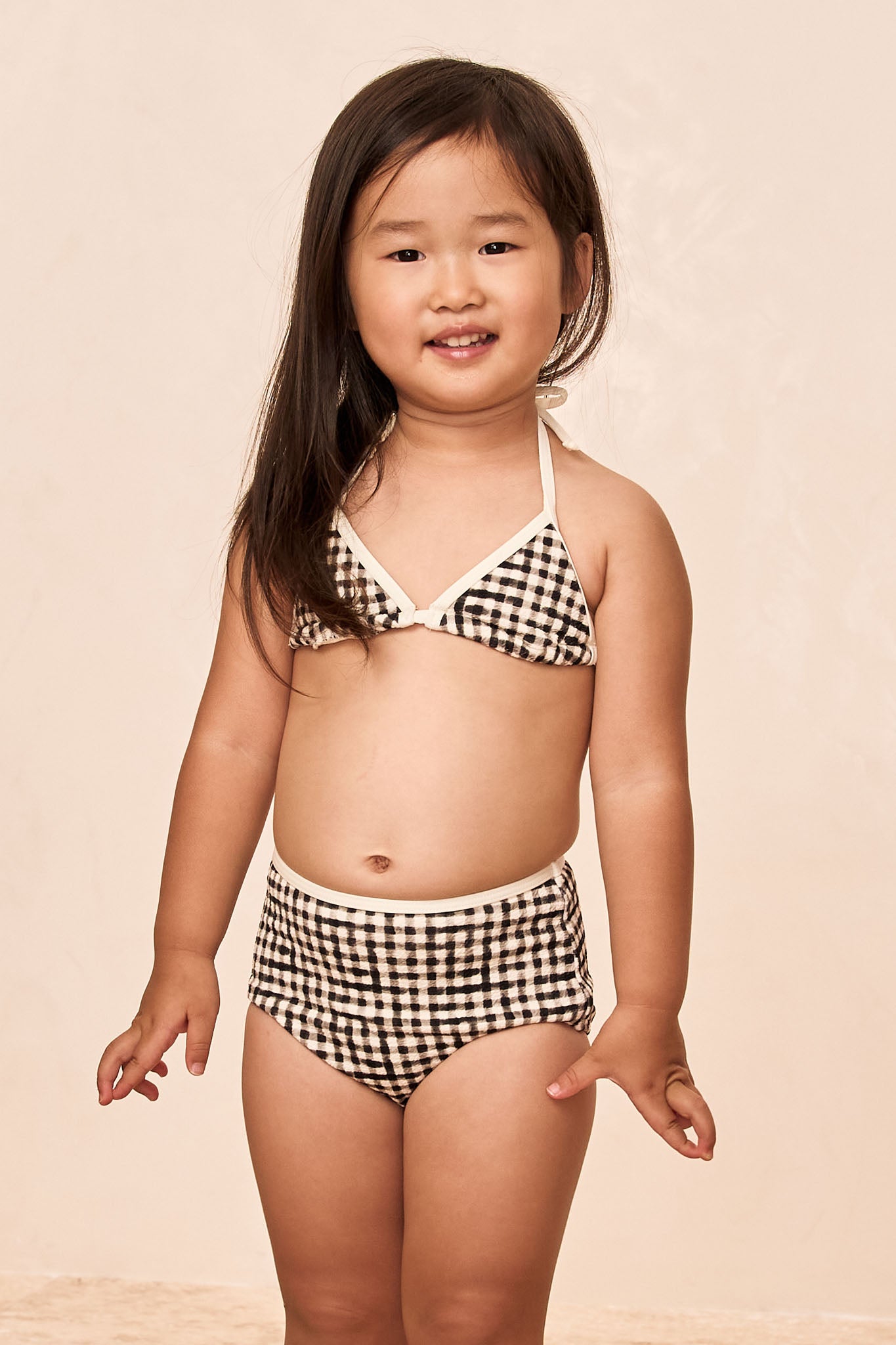 Toddler wearing black-and-white gingham two-piece swimsuit, standing and smiling against a neutral background.