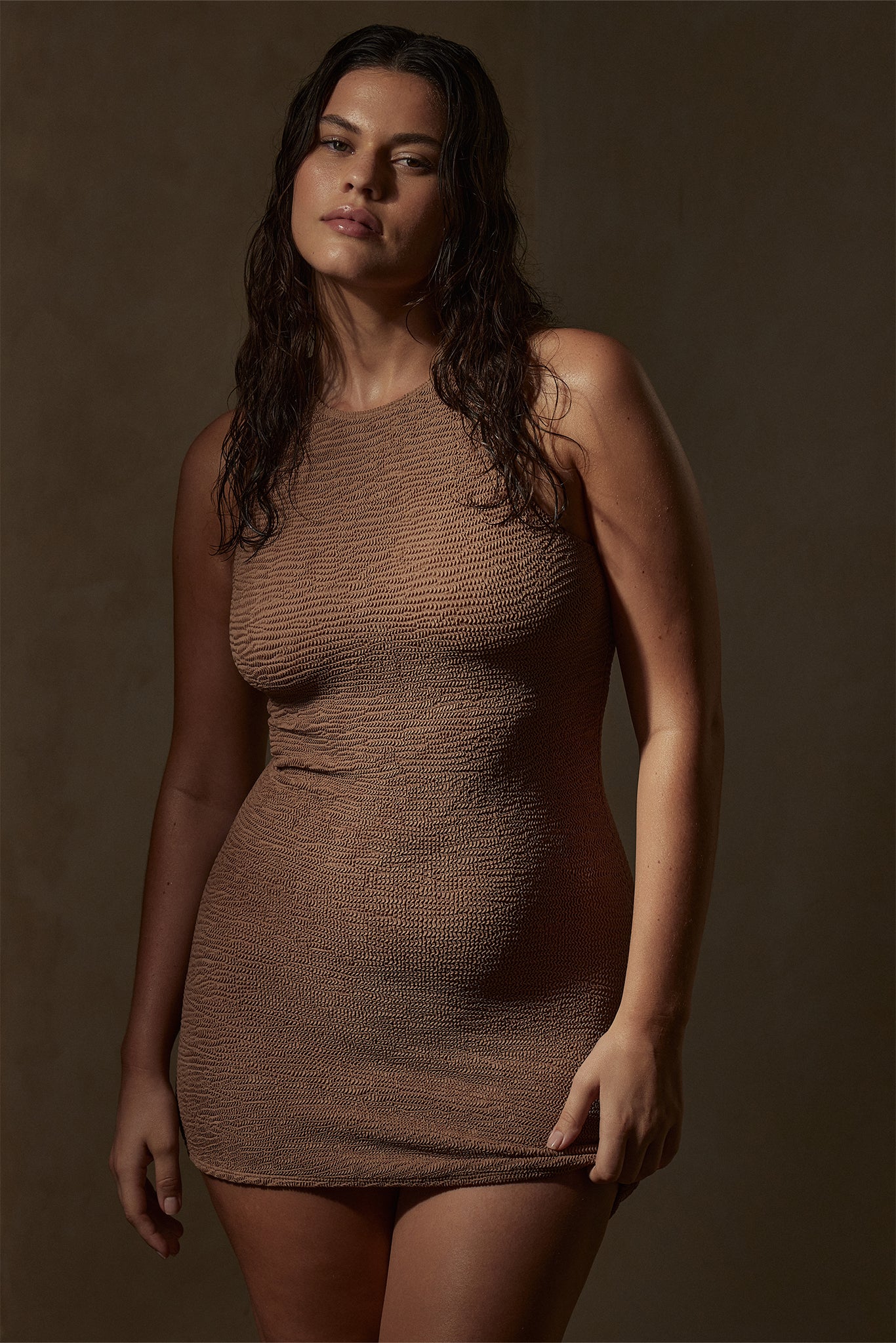 Portrait of a woman with wet hair wearing a fitted brown textured sleeveless knit mini dress on a neutral background.