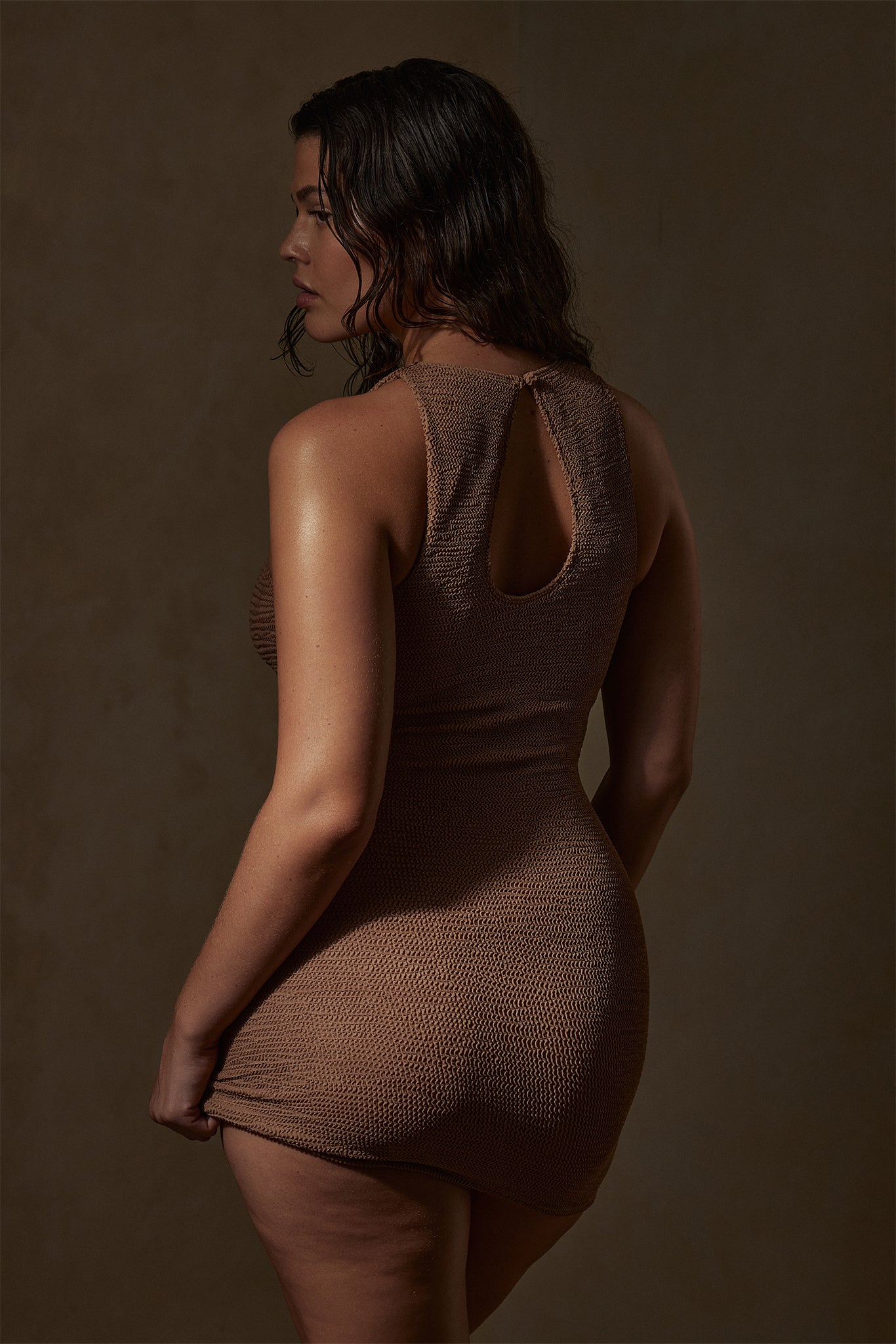 Woman with wet hair, back turned in a sleeveless brown knit mini dress, holding the dress hem.