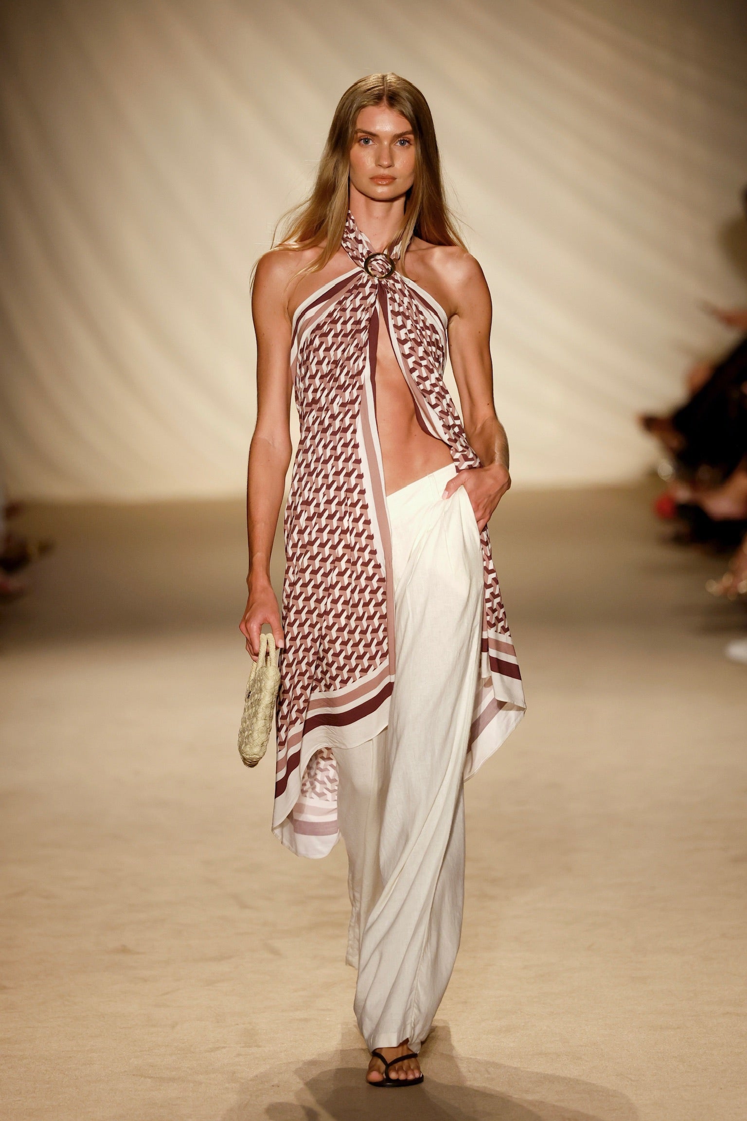 Runway model wearing patterned halter scarf top, wide white pants and sandals, holding a woven clutch on beige catwalk.