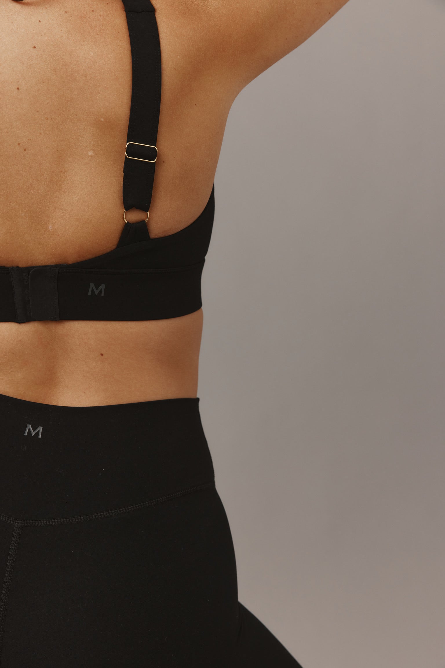 Back view of torso wearing black sports bra and high‑waist leggings, small 'M' logos on bra band and waistband.