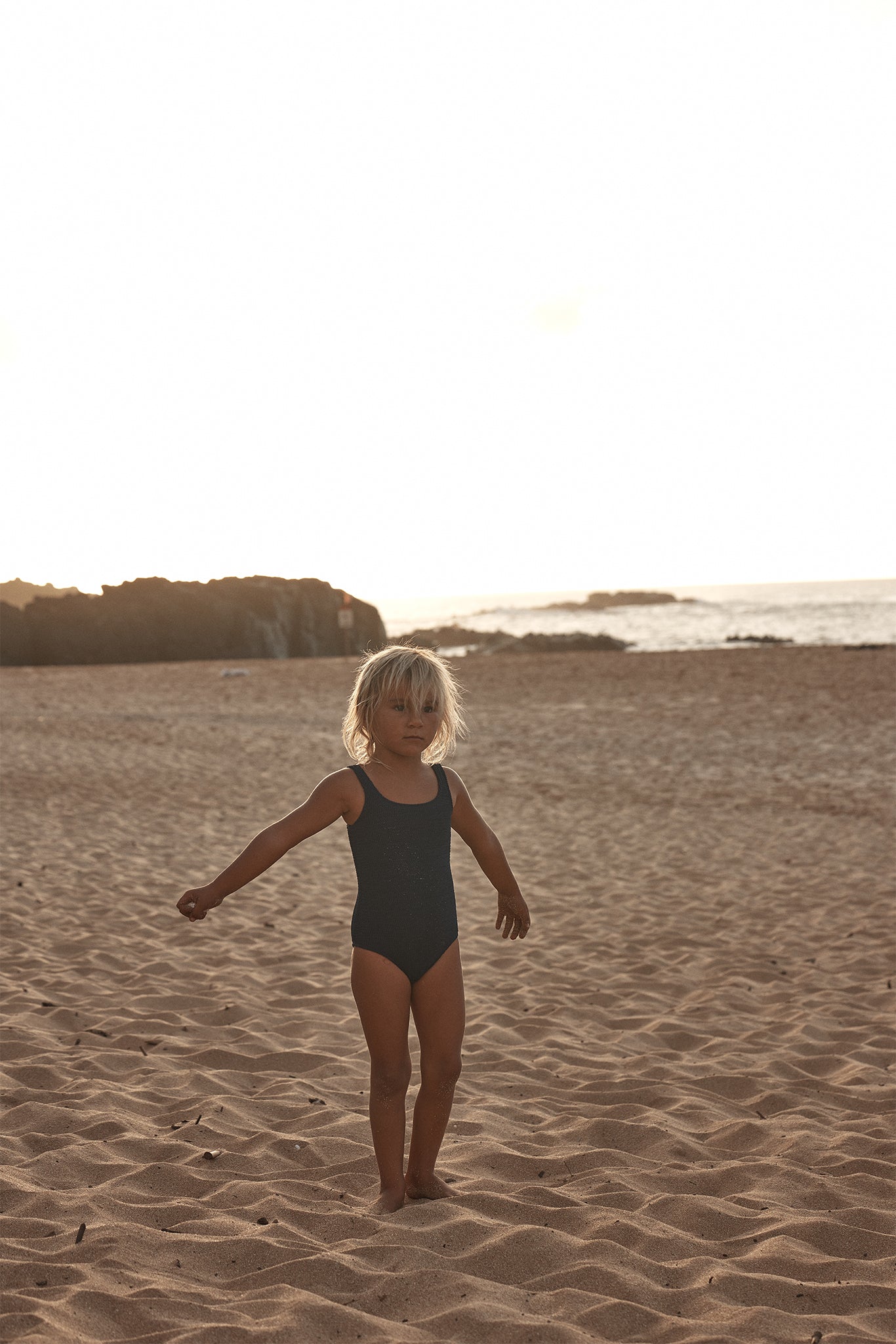 Young child with blond hair standing on sandy beach at sunset wearing a dark one-piece swimsuit