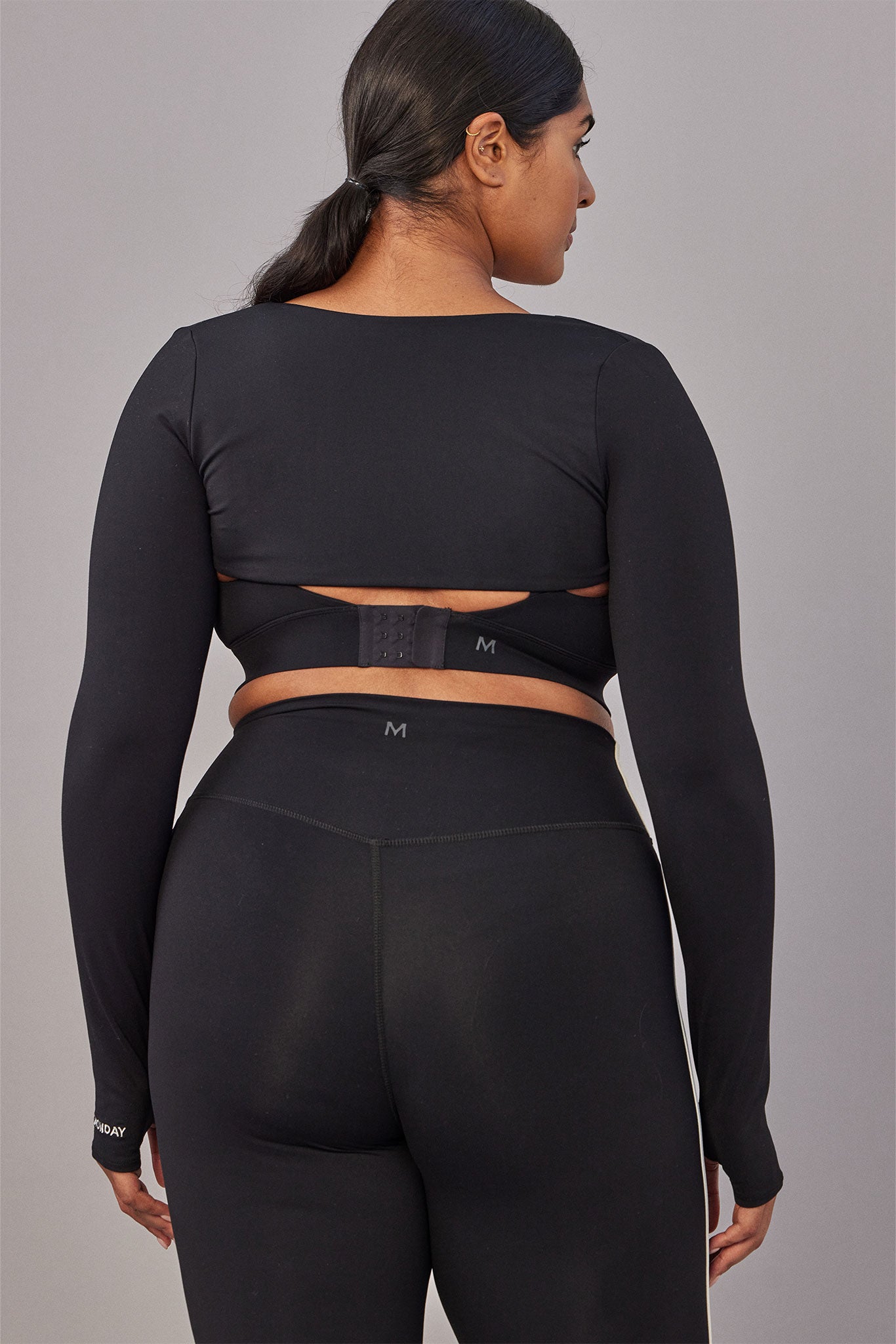 Rear view of person in black long-sleeve crop top and high-waist leggings with 'M' logos and 'MONDAY' on sleeve.